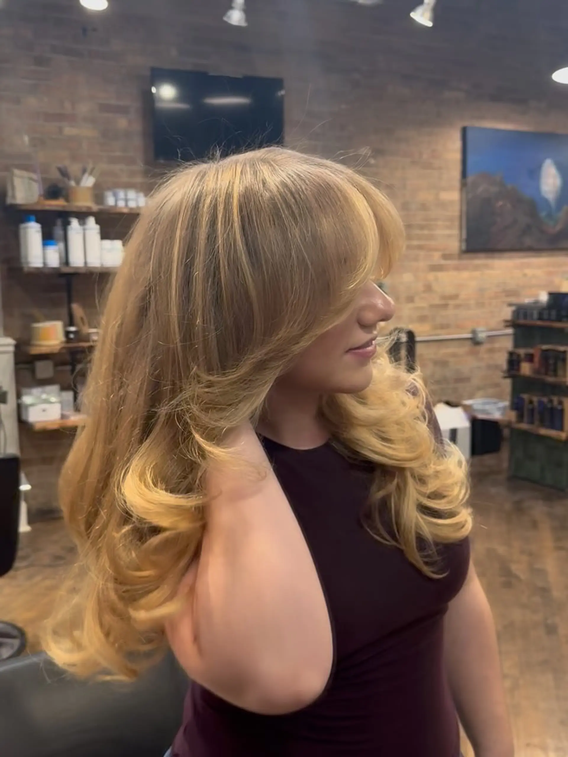 Long warm blonde hair styled in a bouncy, voluminous blowout featuring face-framing curtain bangs and flipped ends. 1