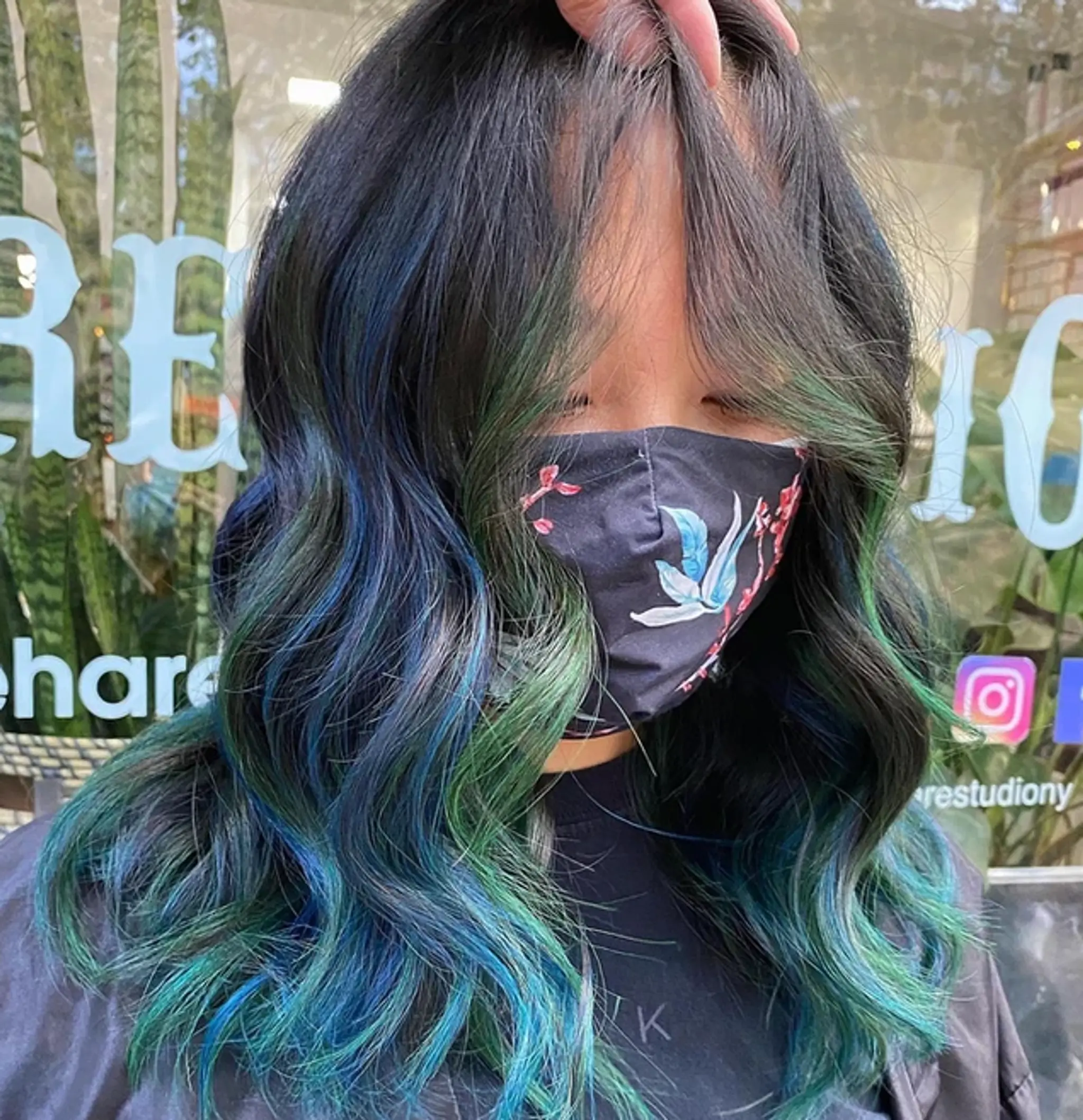 A vibrant medium-length hairstyle with loose waves, featuring a striking mix of black, dark blue, and green hues. The hair is layered to enhance volume and texture.