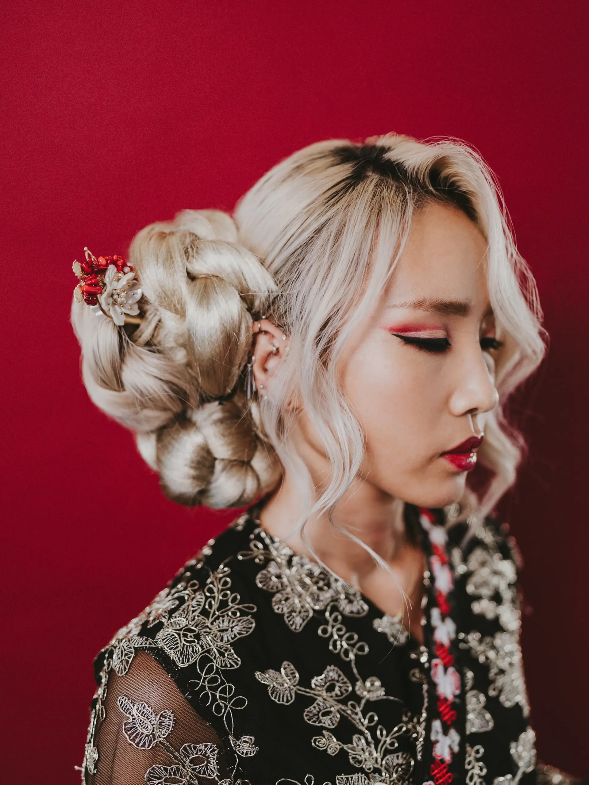 An elaborate, voluminous editorial updo featuring sculptural braided twists in platinum blonde with face-framing tendrils and a traditional floral hairpin. 1