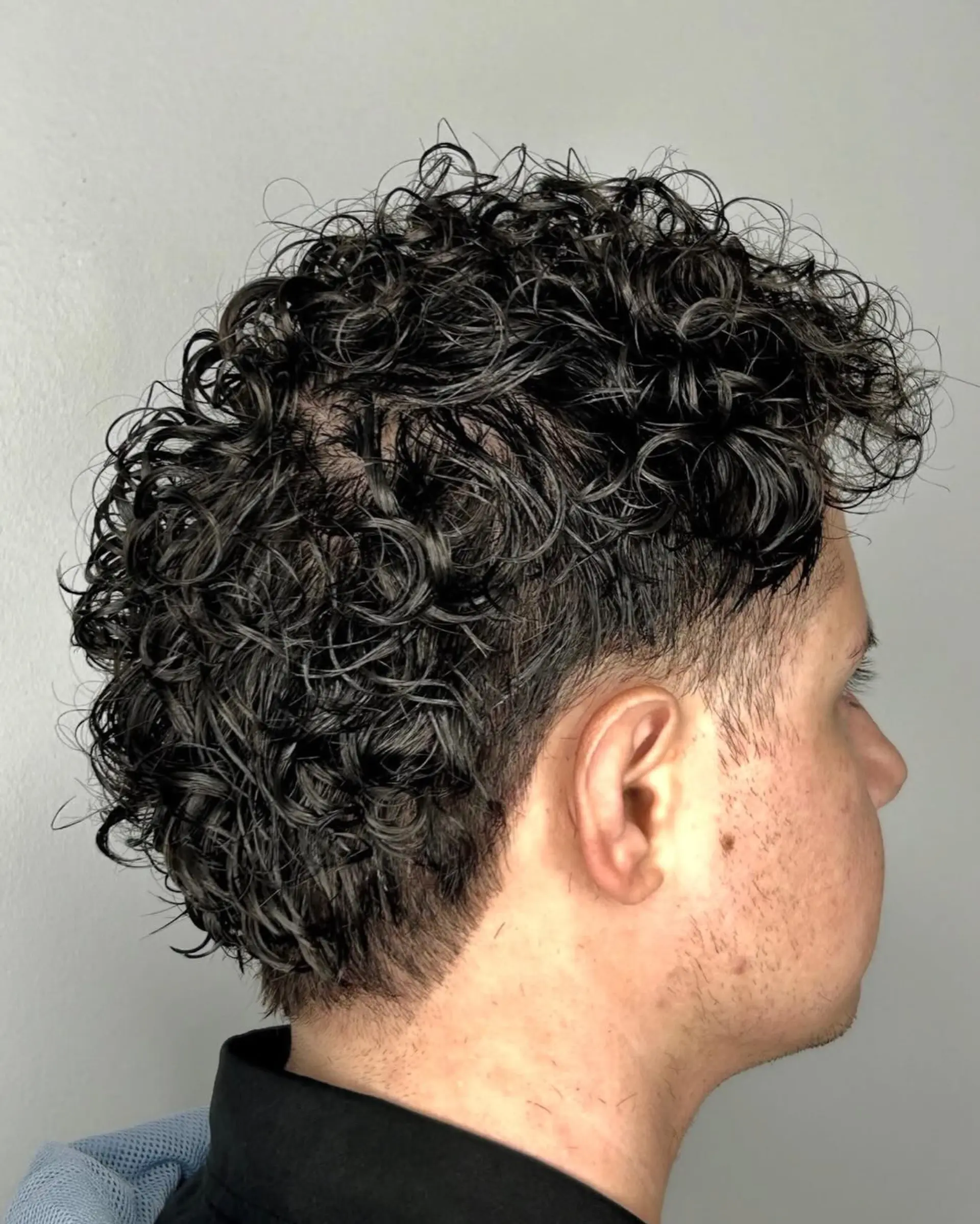 A short men's perm featuring defined, glossy curls styled with natural volume and a clean taper fade around the hairline. 1