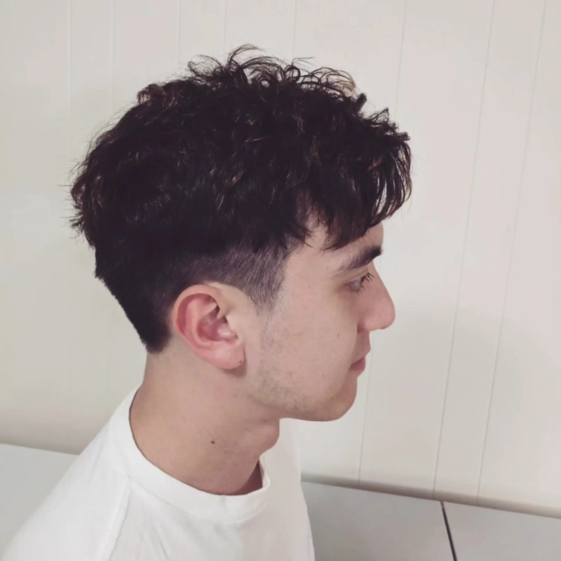 A men's short two-block haircut featuring textured perm curls on top with tapered sides and nape. 1