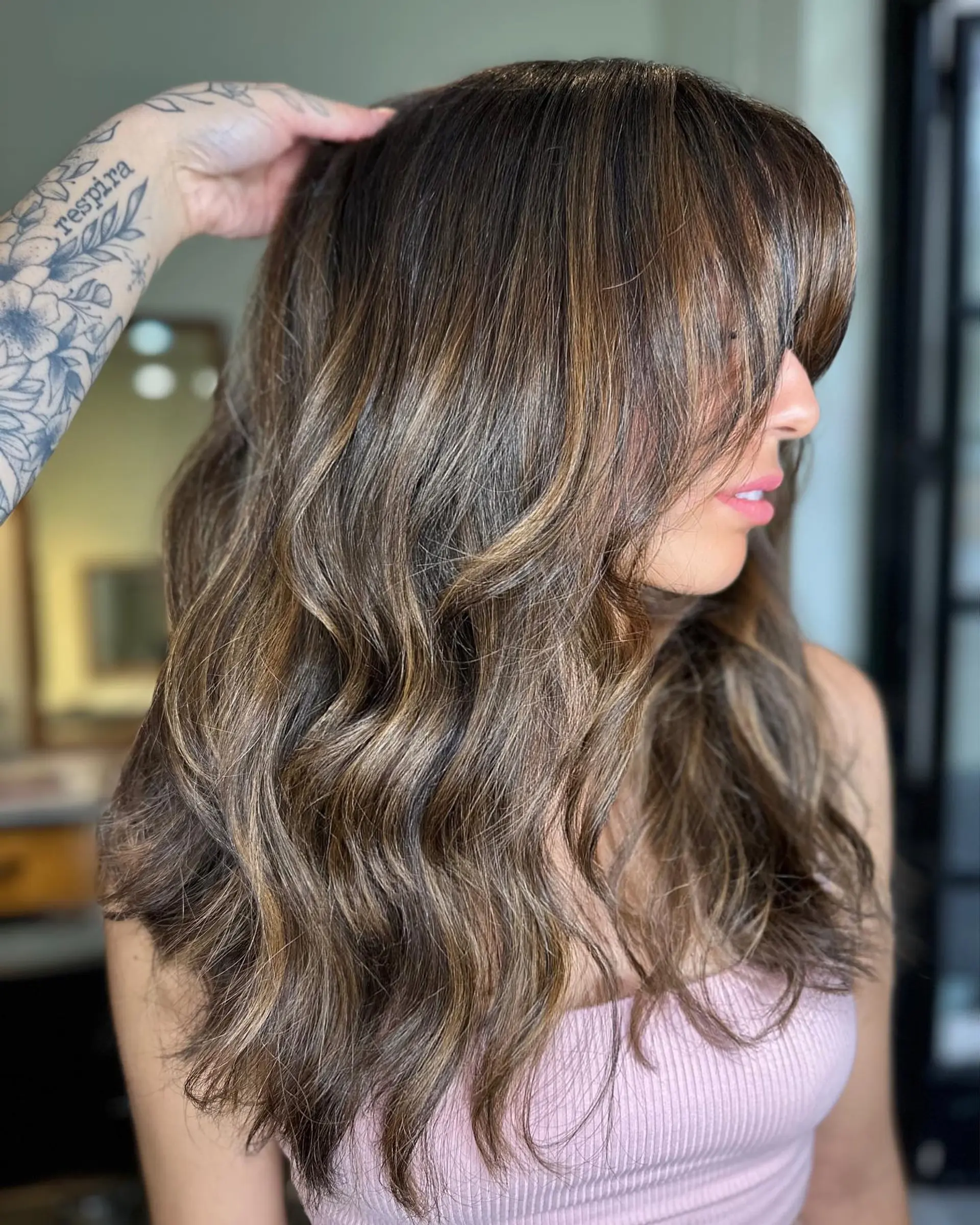 Long layered brunette hair styled in soft waves with warm caramel highlights and face-framing bangs. 1