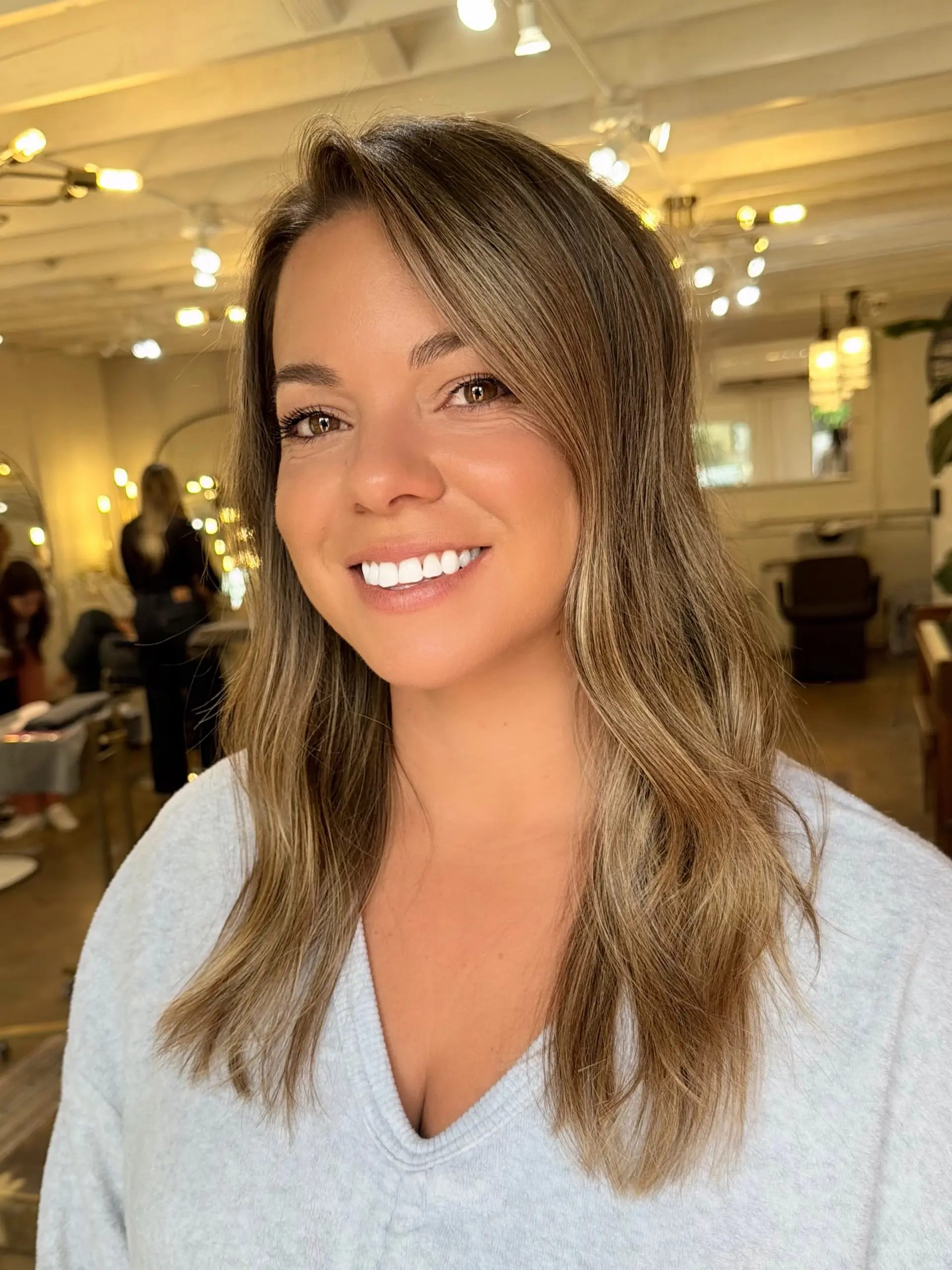 Sun-kissed bronde highlights blended into a natural brown base, styled with soft, smooth waves and a side part. 1