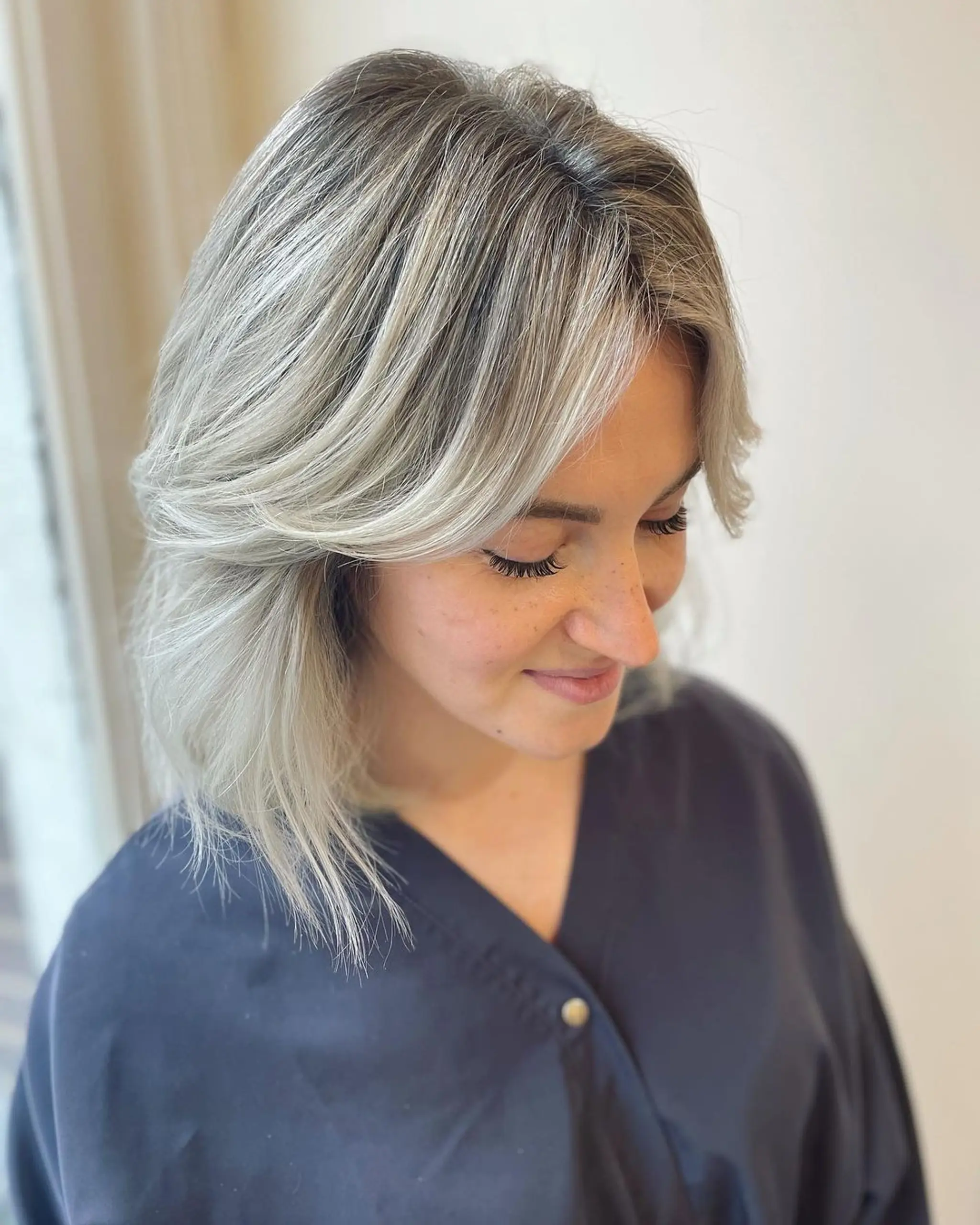 Medium-length wavy silver and ash blonde hair with soft layers and a side part, featuring feathered ends for a textured look.