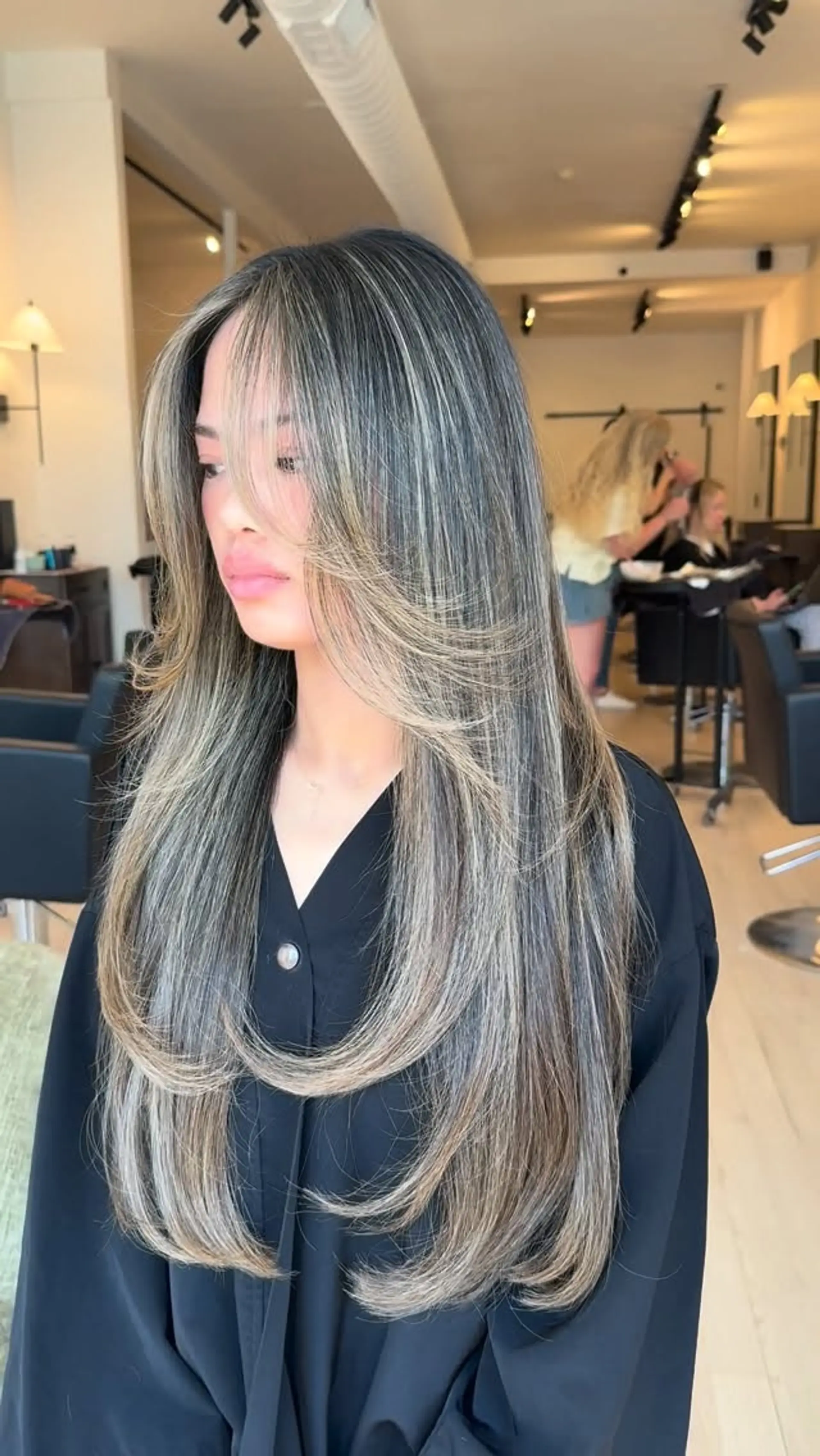 Long dark hair styled with heavy dimensional ash blonde highlights, face-framing curtain fringe, and smoothly curled-under layered ends. 1
