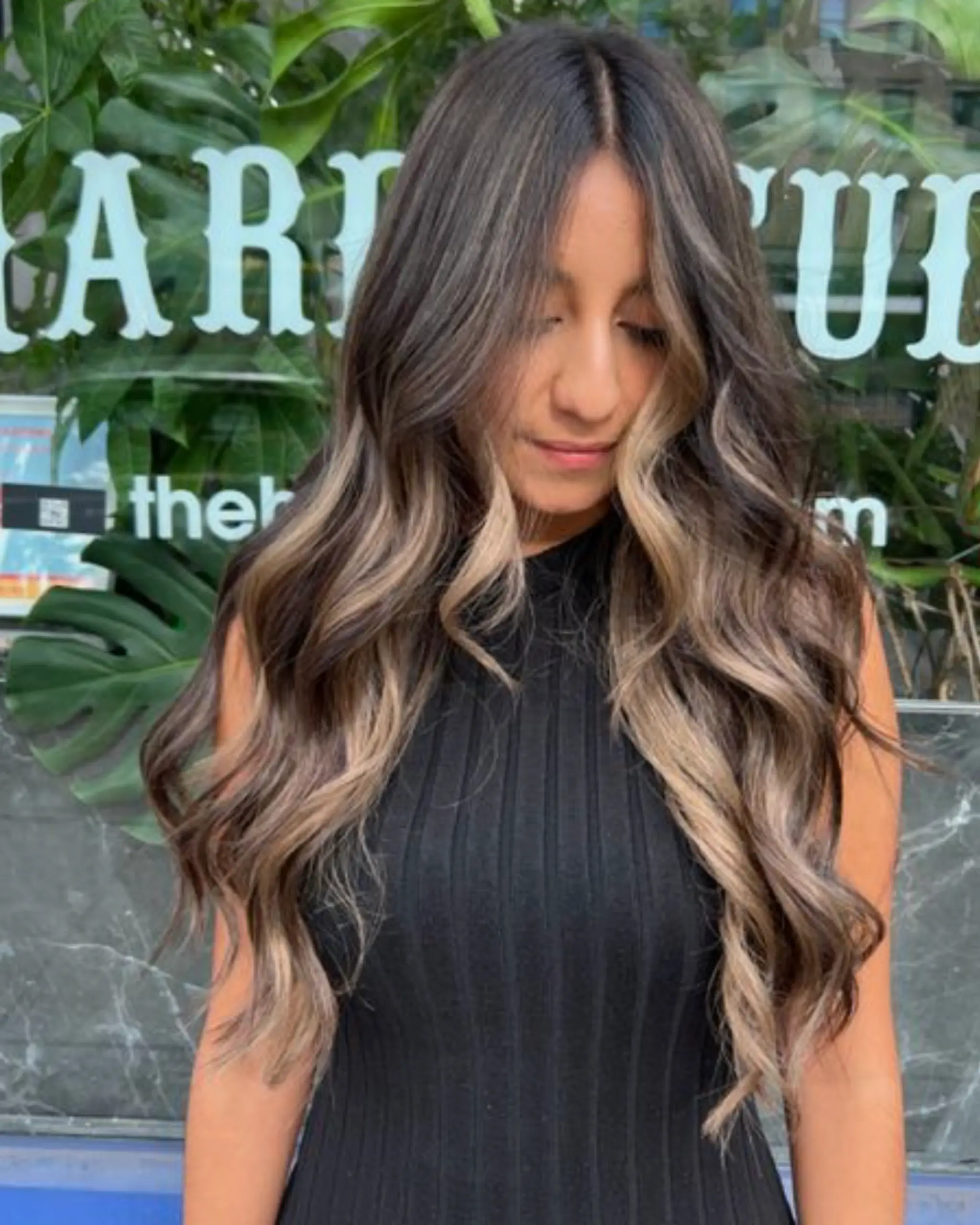 Long, wavy locks with a natural dark brown base expertly blended with blonde highlights creating a stunning balayage effect. Center part with layered waves.