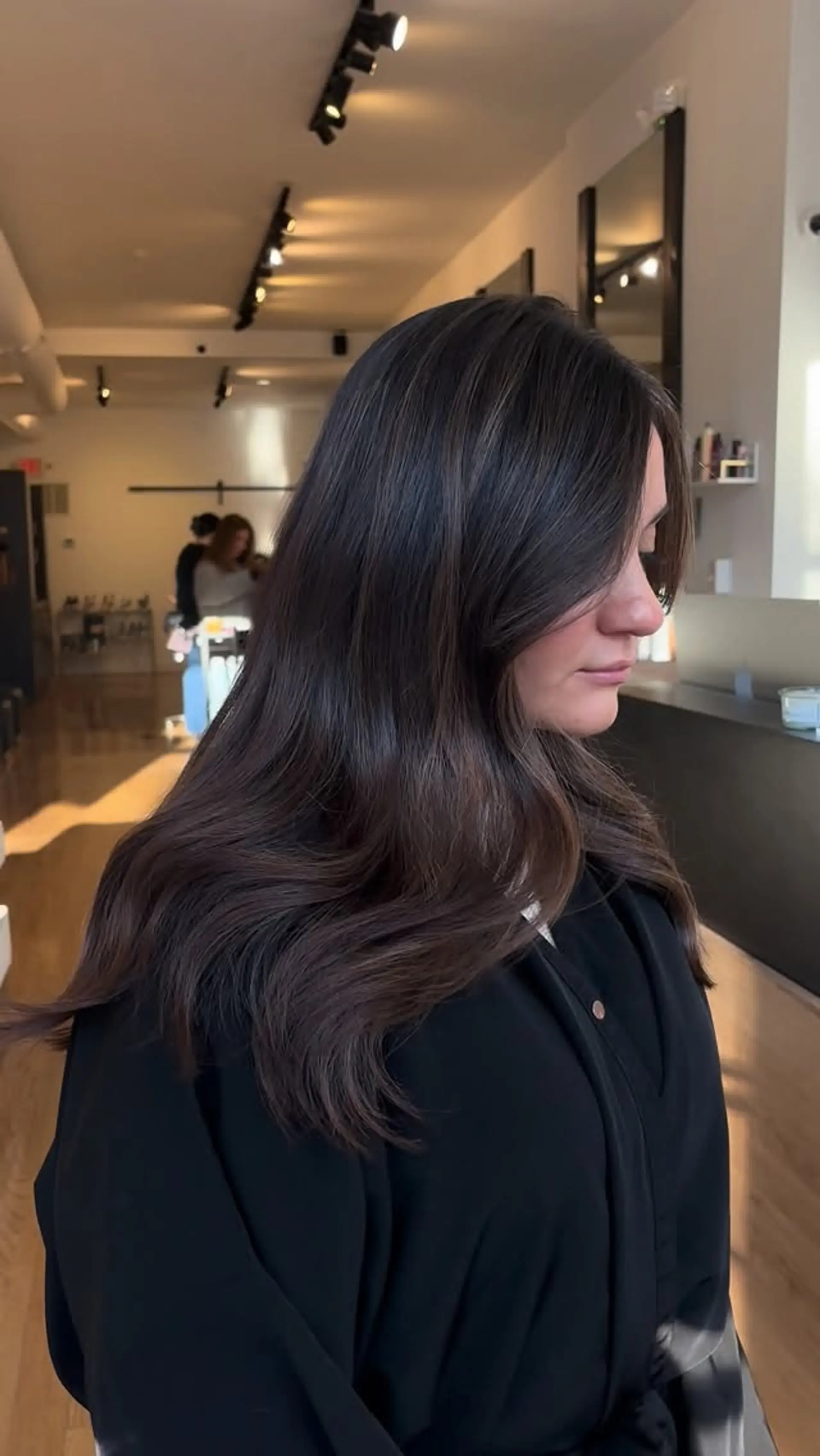 A glossy, rich chocolate brunette style featuring subtle dimension and soft, bouncy blowout waves. 1