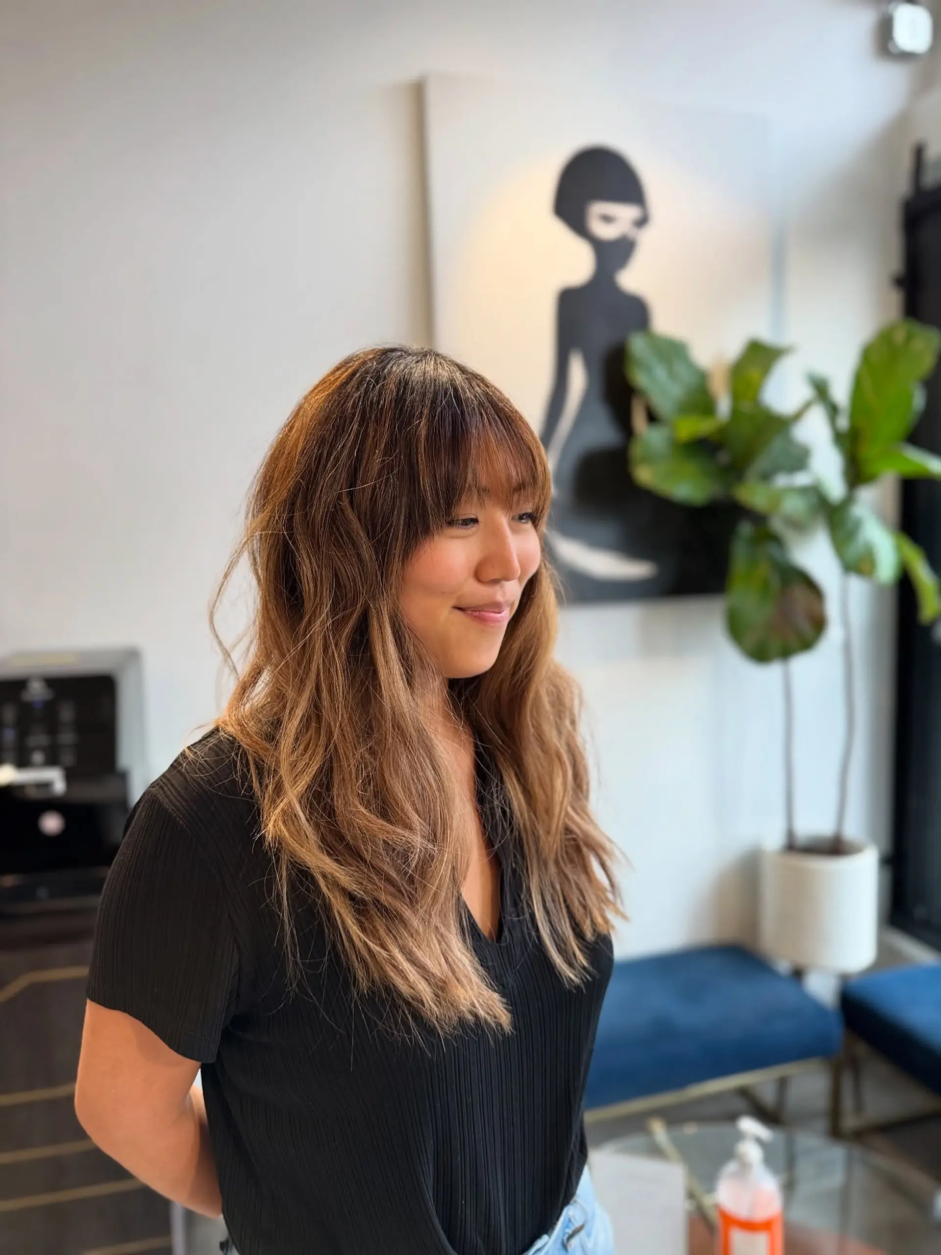 Long layered brunette hair styled in loose tousled waves with straight-across textured bangs and subtle caramel balayage. 1