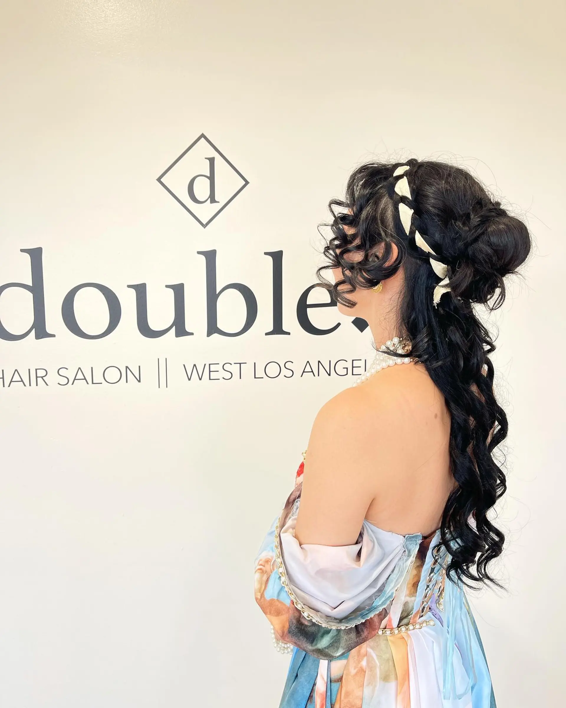 An intricate, romantic half-up hairstyle featuring tight defined curls cascading down the back, accented with an interwoven light-colored ribbon around the crown and soft tendrils framing the face. 1