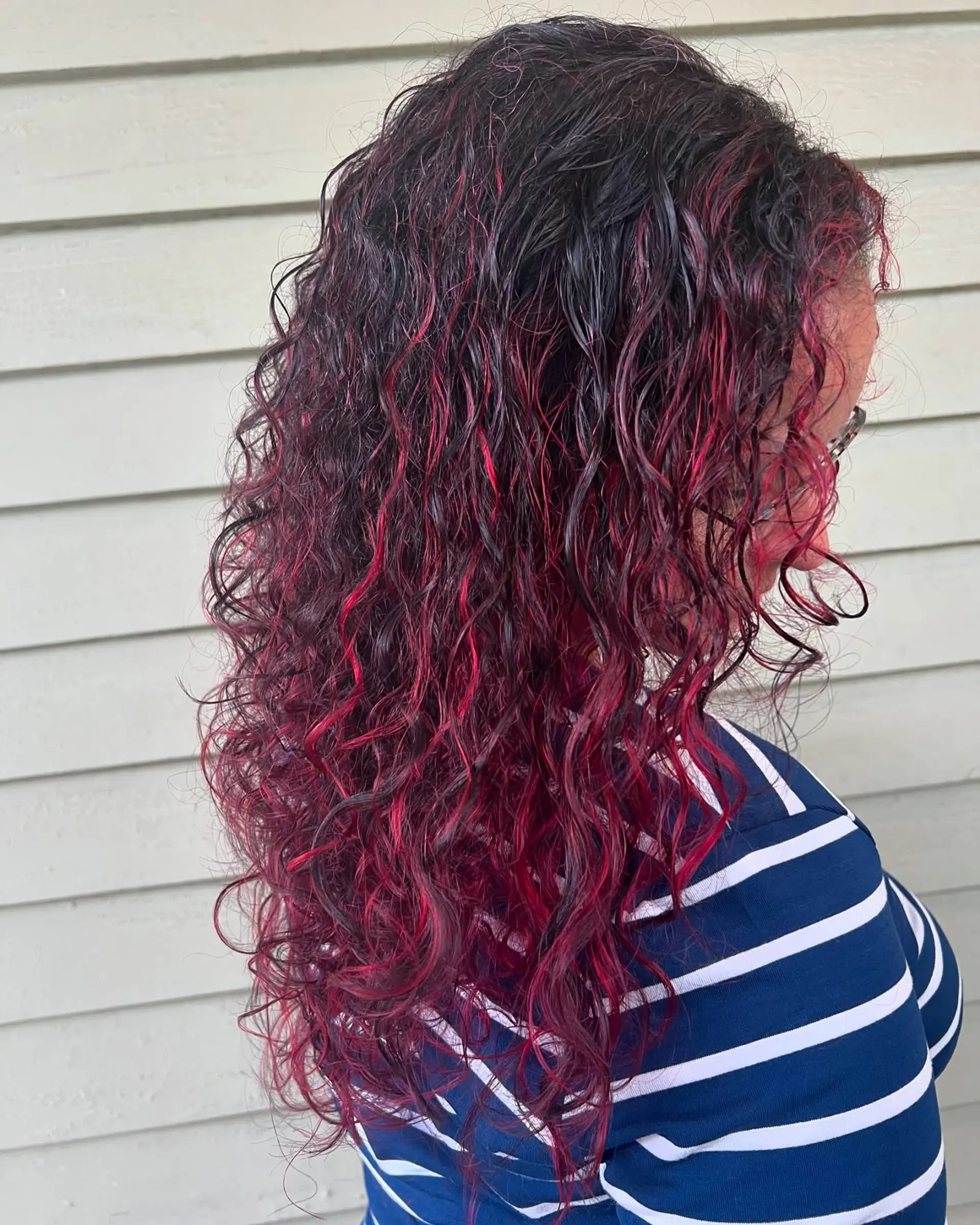Long curly hair styled with a vivid cherry red or magenta balayage, featuring a deep, dark root that transitions into bright, playful curls for a dynamic and textured look. 1