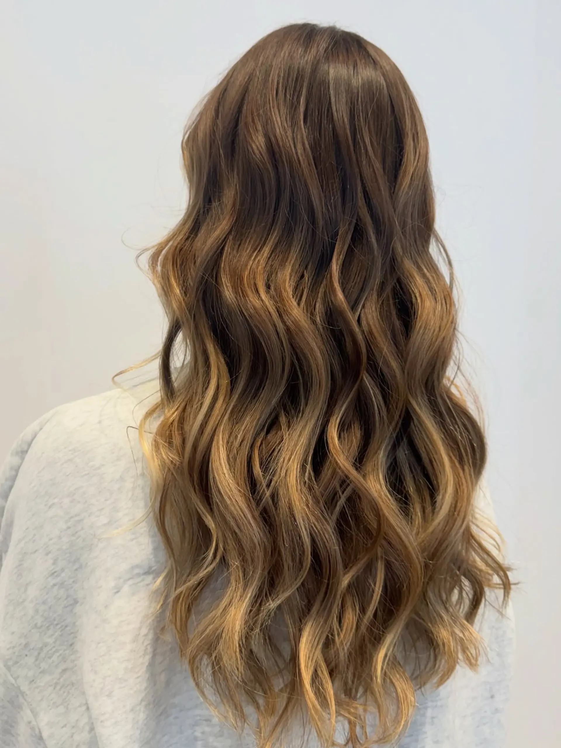 Long wavy brunette hair enhanced with warm caramel and honey balayage highlights. 1