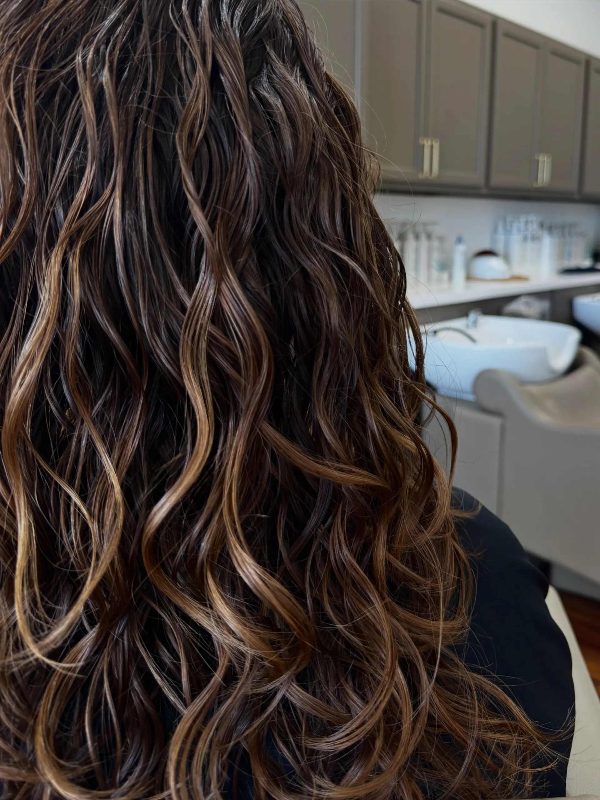 Long brunette hair styled with defined, glossy natural waves and loose ringlets featuring soft caramel highlights. 1