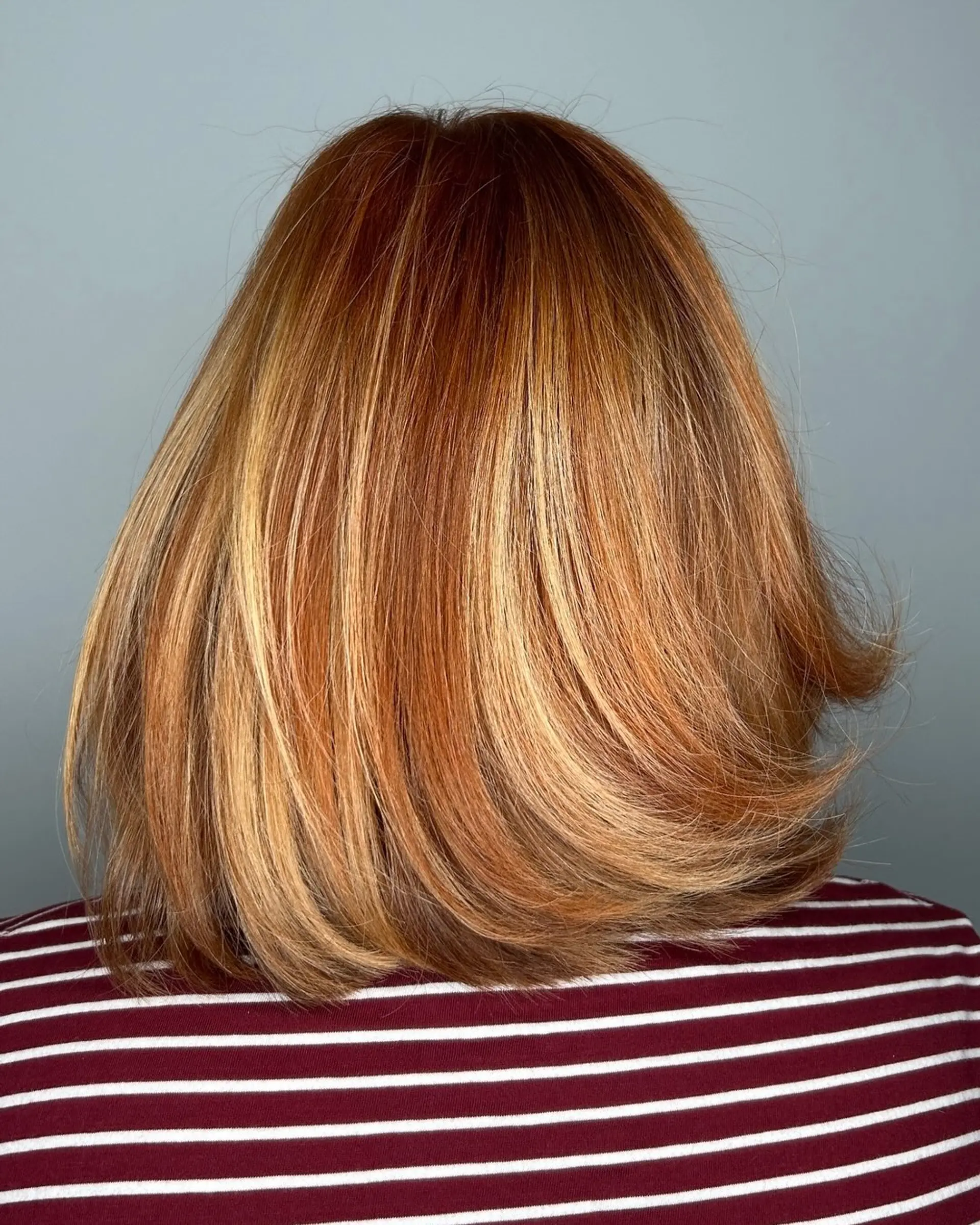 A medium-length layered lob featuring dimensional copper and golden blonde tones with curled-under ends. 1