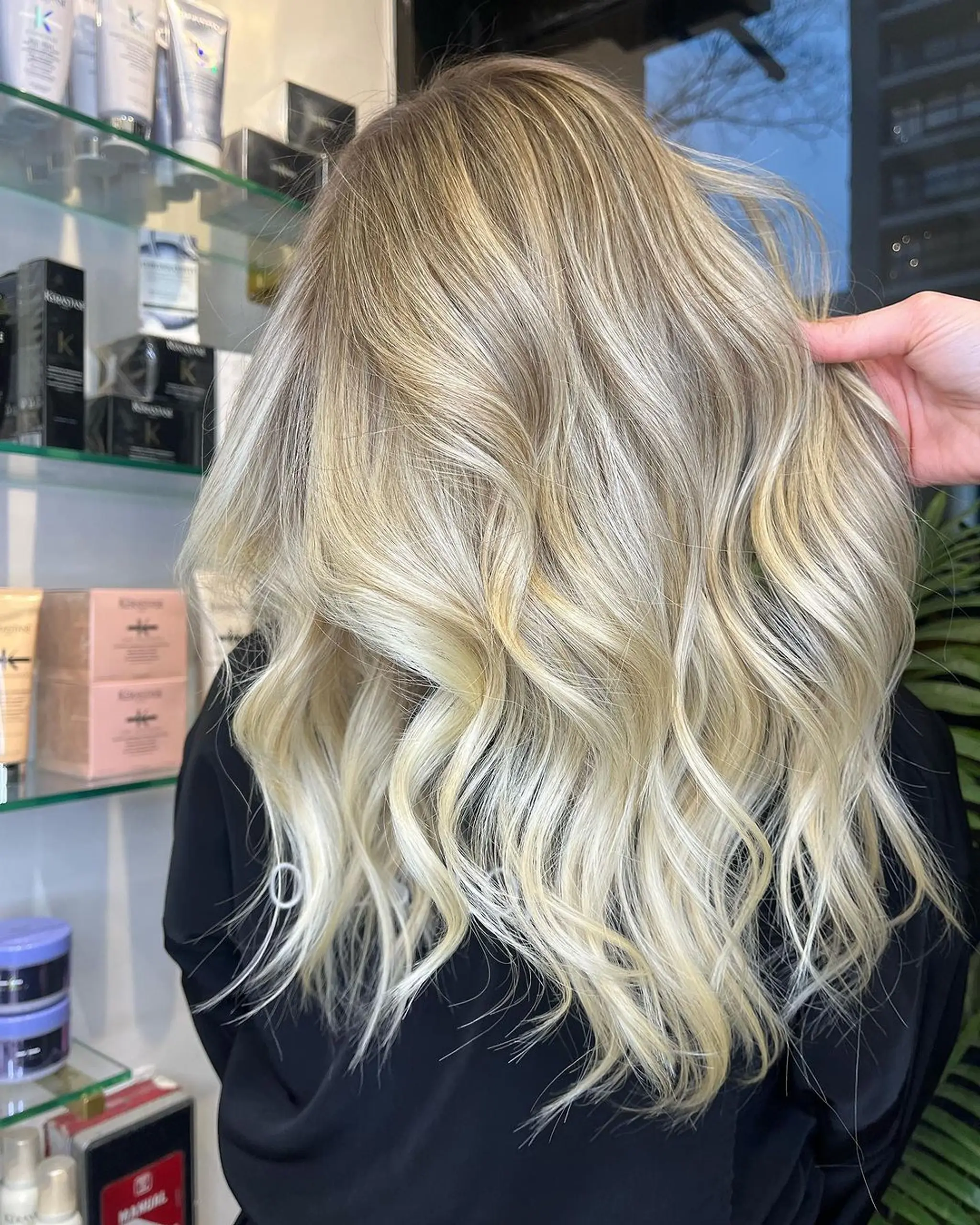 This hairstyle features long, wavy hair with a balayage that transitions from a deep blonde at the roots to a platinum blonde at the ends, creating a natural sun-kissed effect. The waves are soft and loose, enhancing the layered cut that adds volume and movement.