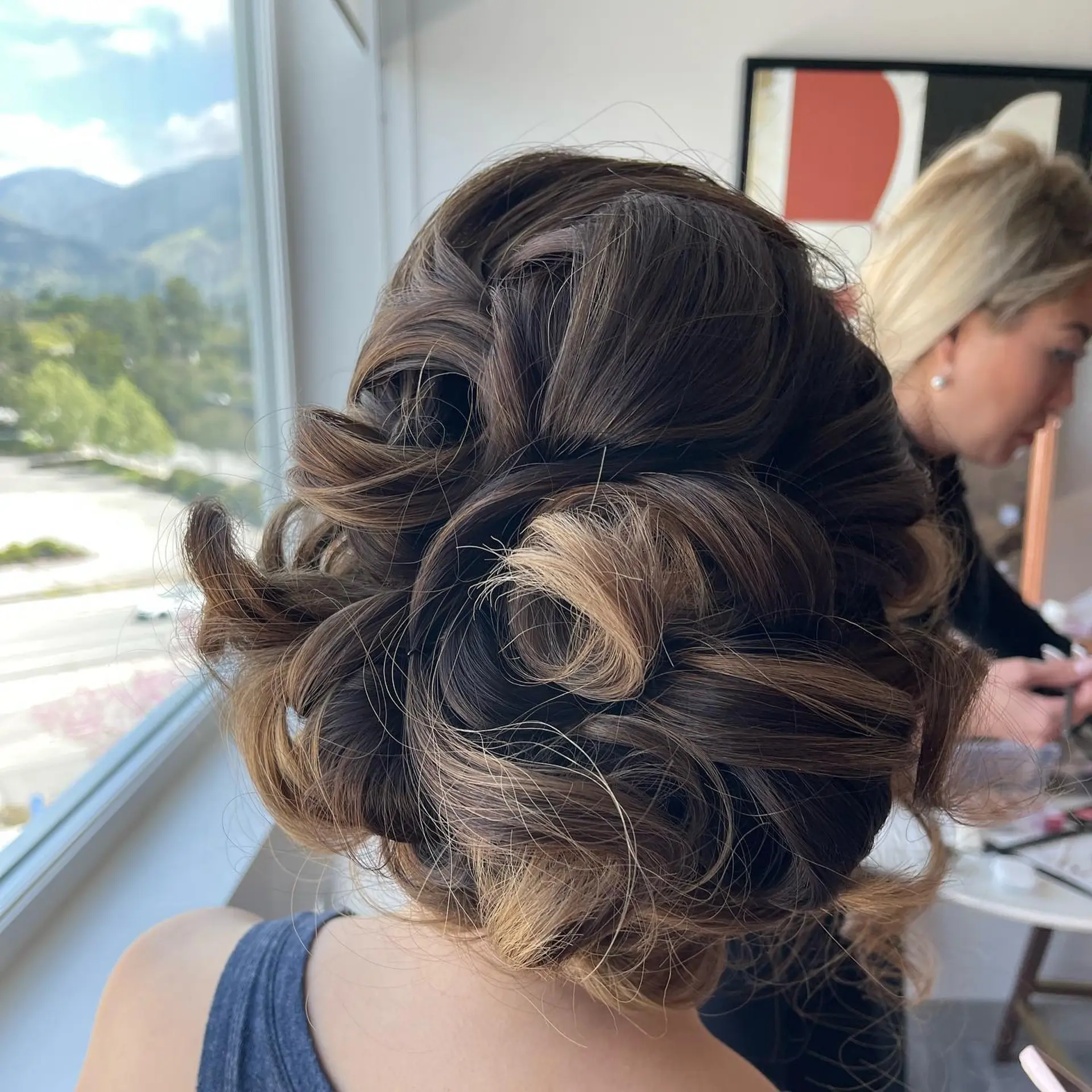 A textured low bun upstyle with soft loops, pinned curls, and dimensional caramel highlights. 1