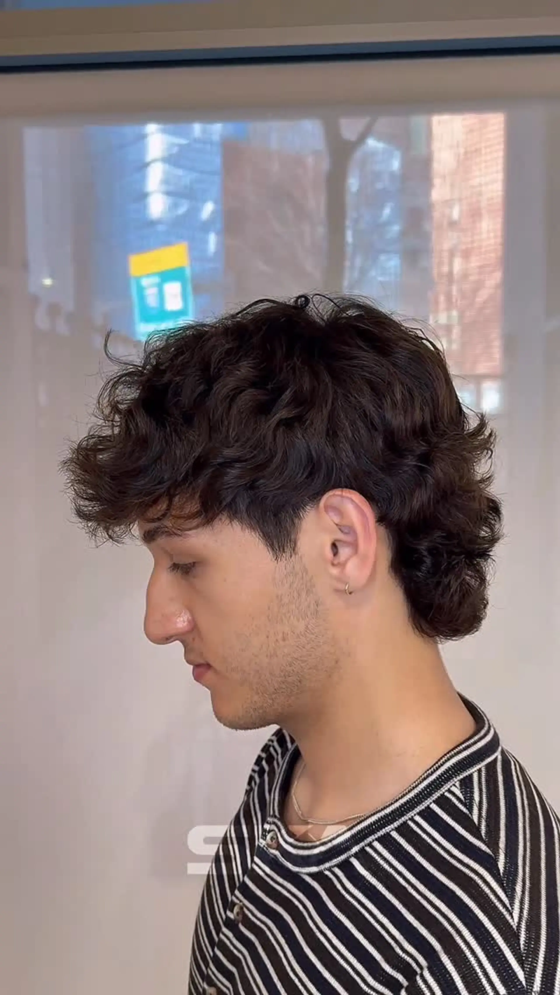 A modern wavy mullet haircut with textured curls on top, neat tapered sides around the ear, and layered length at the nape. 1