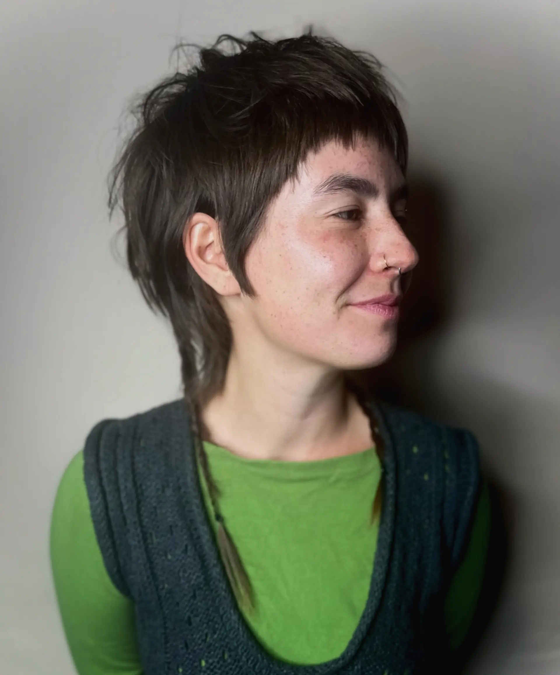 A playful and edgy mixie haircut combining elements of a pixie and a mullet. It features blunt textured micro bangs, soft sideburn detailing, and layered, tousled volume through the crown and nape. 1