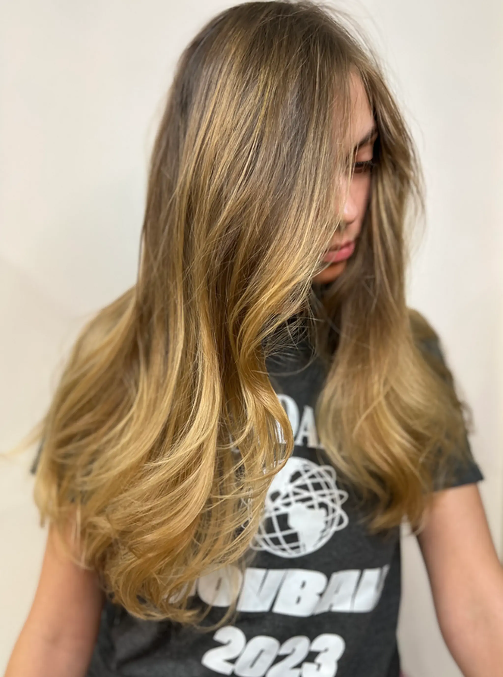 Long wavy hairstyle with face-framing layers and a blend of dark and light blonde highlights, creating a sunlit effect.