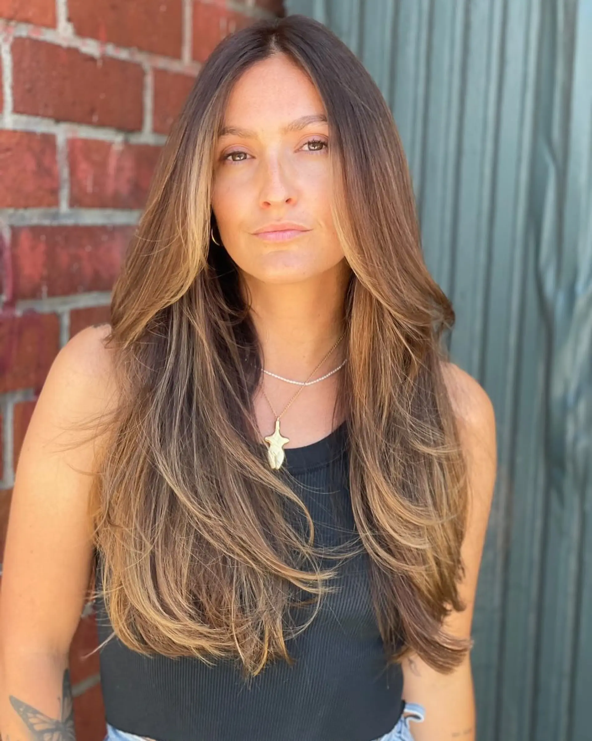 Long brunette hair cut in cascading face-framing layers with sun-kissed warm highlights and a smooth blowout. 1