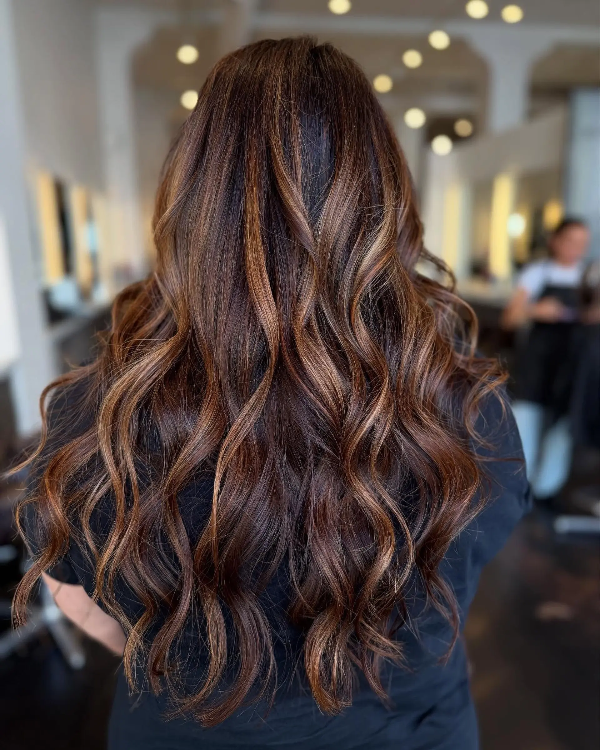 Long brunette hair styled in loose cascading waves with warm caramel balayage highlights. 1
