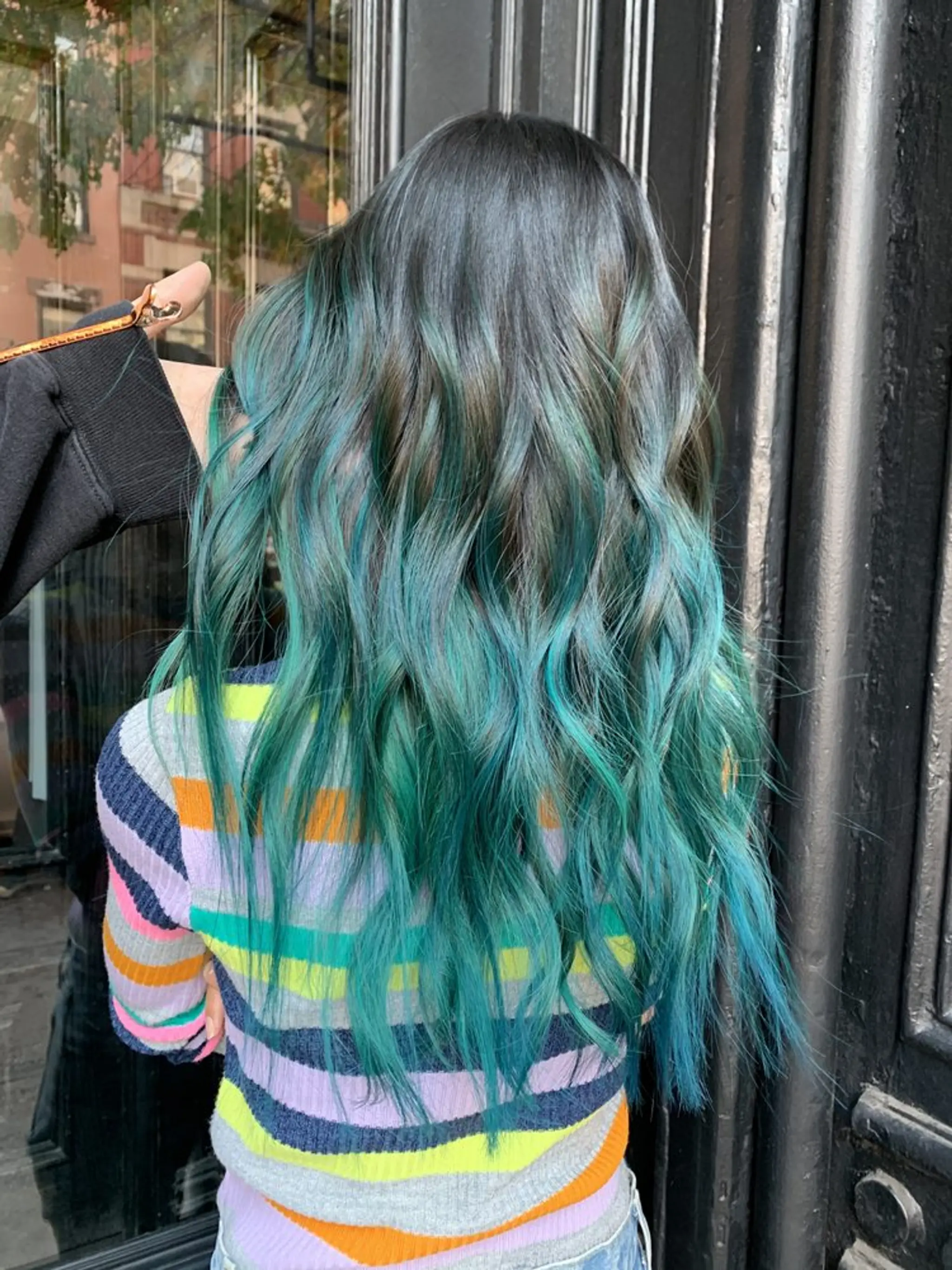 A vibrant ombre hairstyle transitioning from black to teal and dark green. The wavy, long locks are layered to add depth and volume, styled to resemble beach waves.
