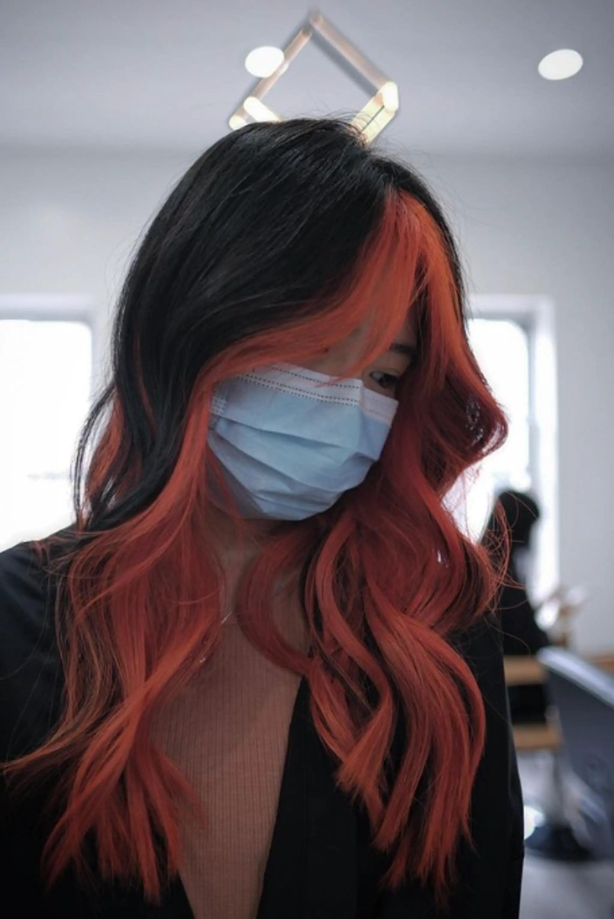 Wavy, medium-length hair with a two-tone color scheme of black and vibrant orange-red. Loose waves and face-framing highlights enhance the layered look.