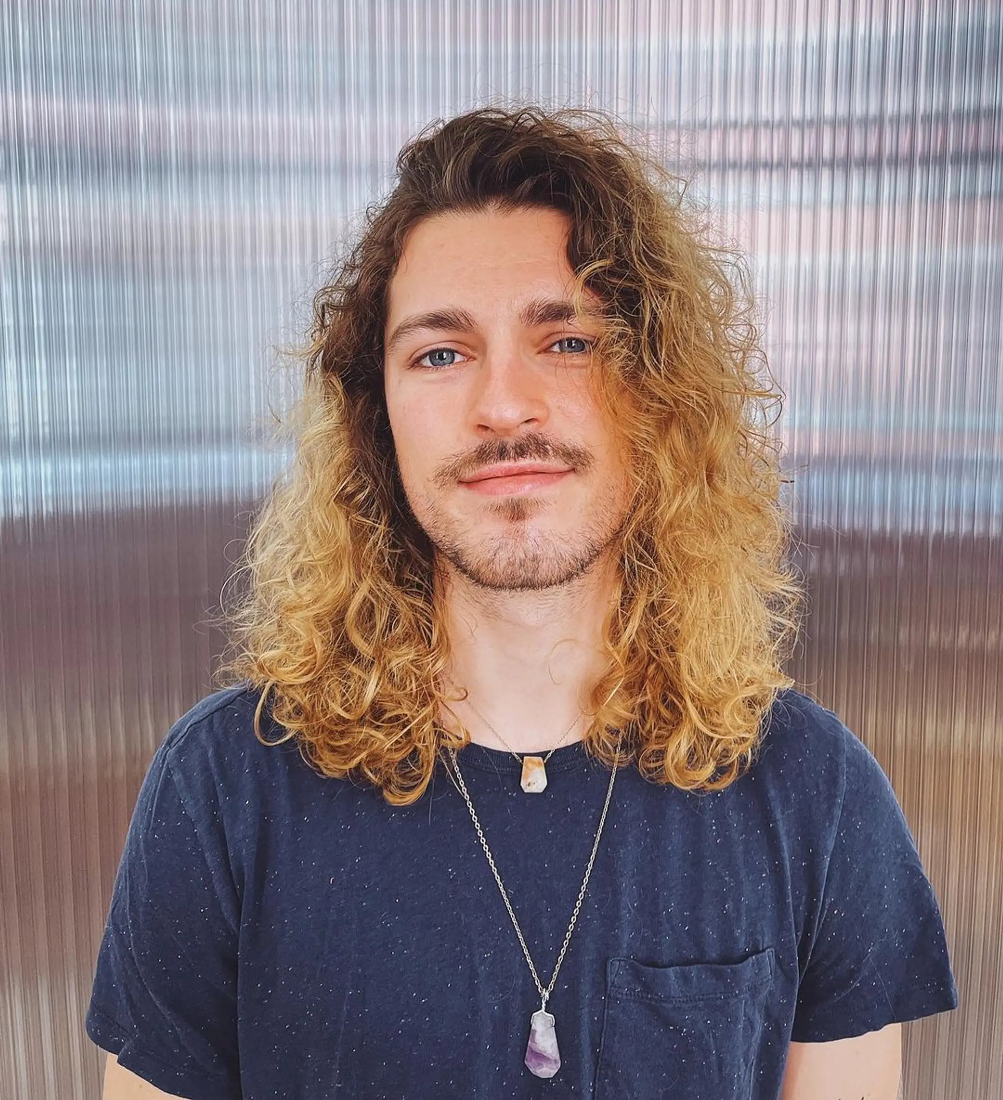 Medium-length, naturally curly hair with a middle part and an ombre effect, transitioning from darker brown roots to golden blonde ends. The curls are loose and voluminous.