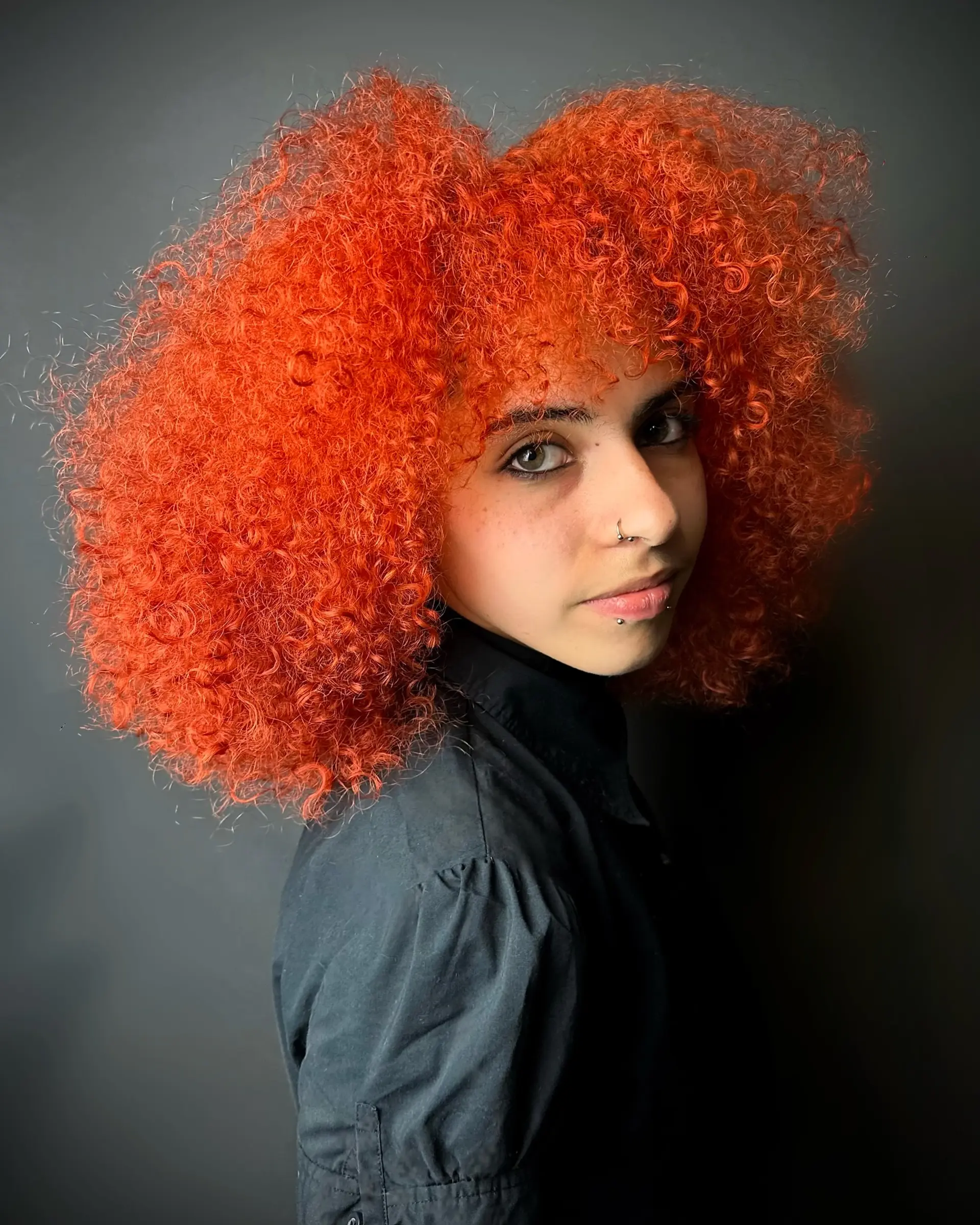 Voluminous mid-length cut on tight, vibrant neon-orange curls with full curly bangs. 1