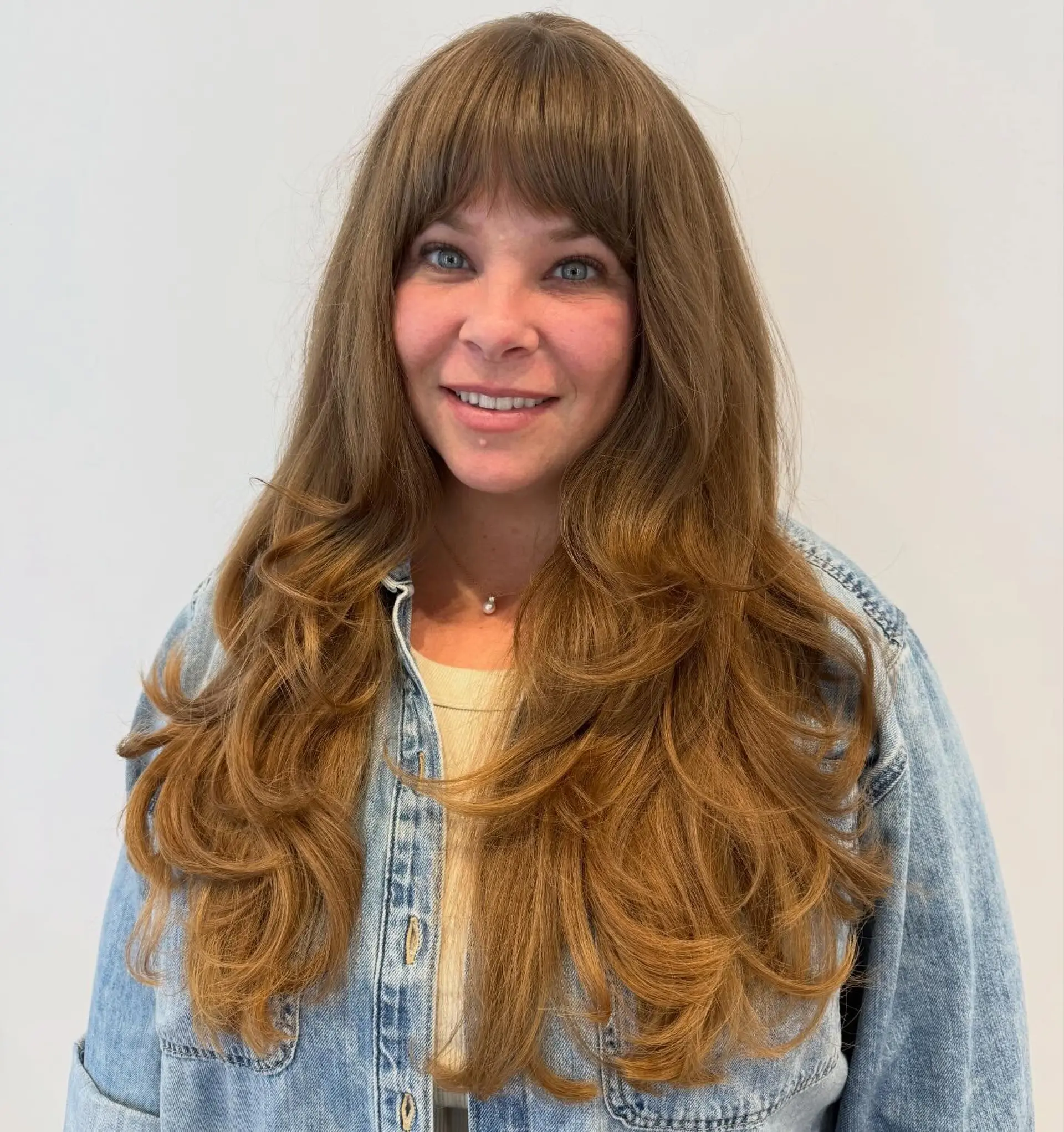 Long warm brown hair cut into voluminous face-framing layers with full straight-across fringe bangs. 1