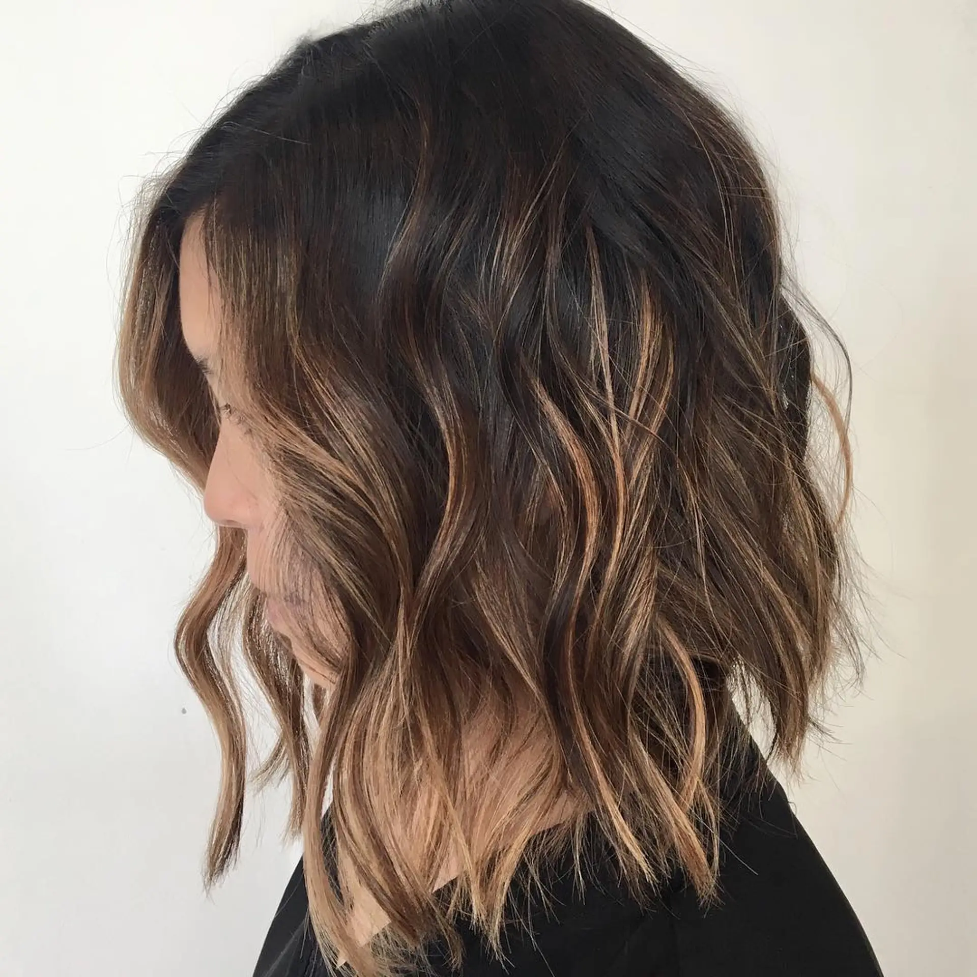 A textured, collarbone-length lob styled with loose beach waves and warm caramel foilayage highlights over a dark espresso base. 1