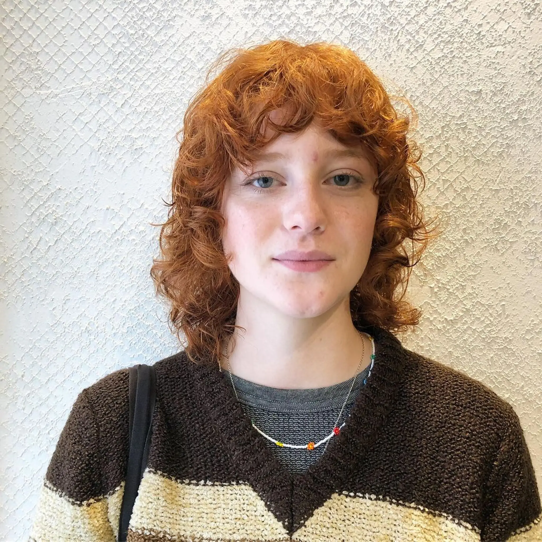 A medium-length curly hairstyle in vibrant auburn and copper tones, featuring soft bangs and a natural layered look.