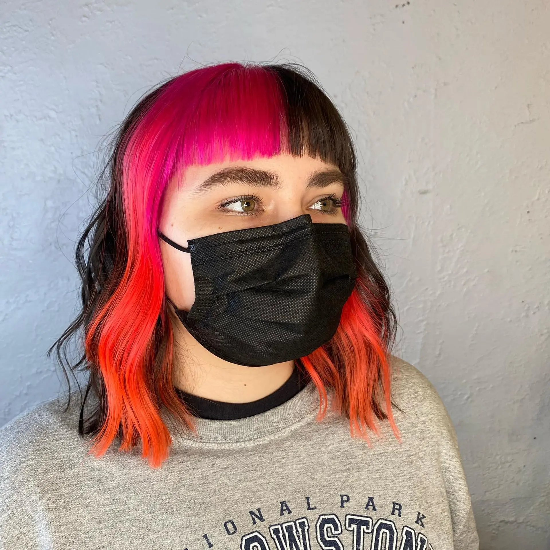 A wavy lob featuring a split dye with one side vibrant pink and the other side bright orange fading into brown, paired with short, blunt baby bangs. 1