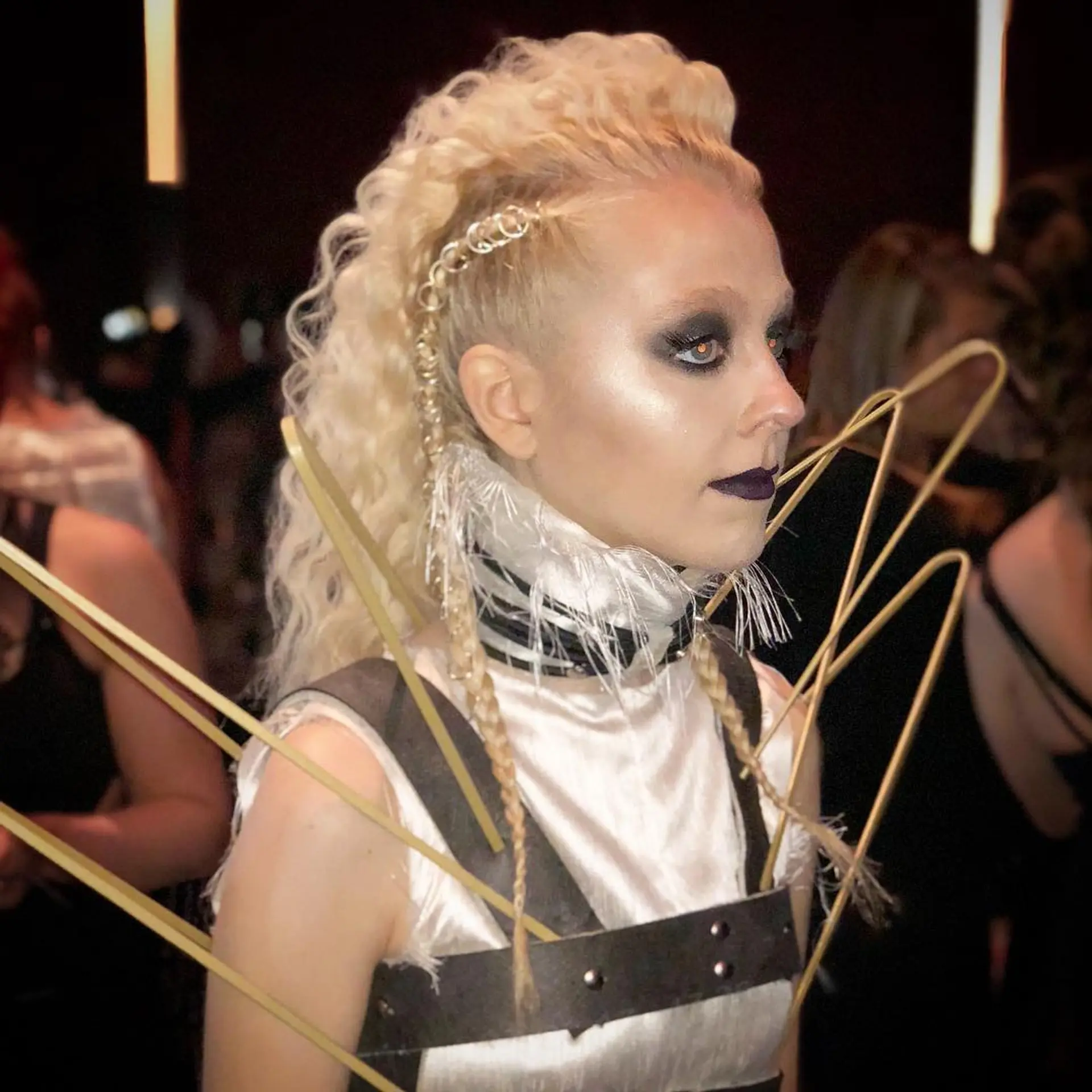 An edgy avant-garde faux hawk featuring voluminous platinum blonde curls, a buzz cut side undercut, and delicate braids embellished with hair rings. 1