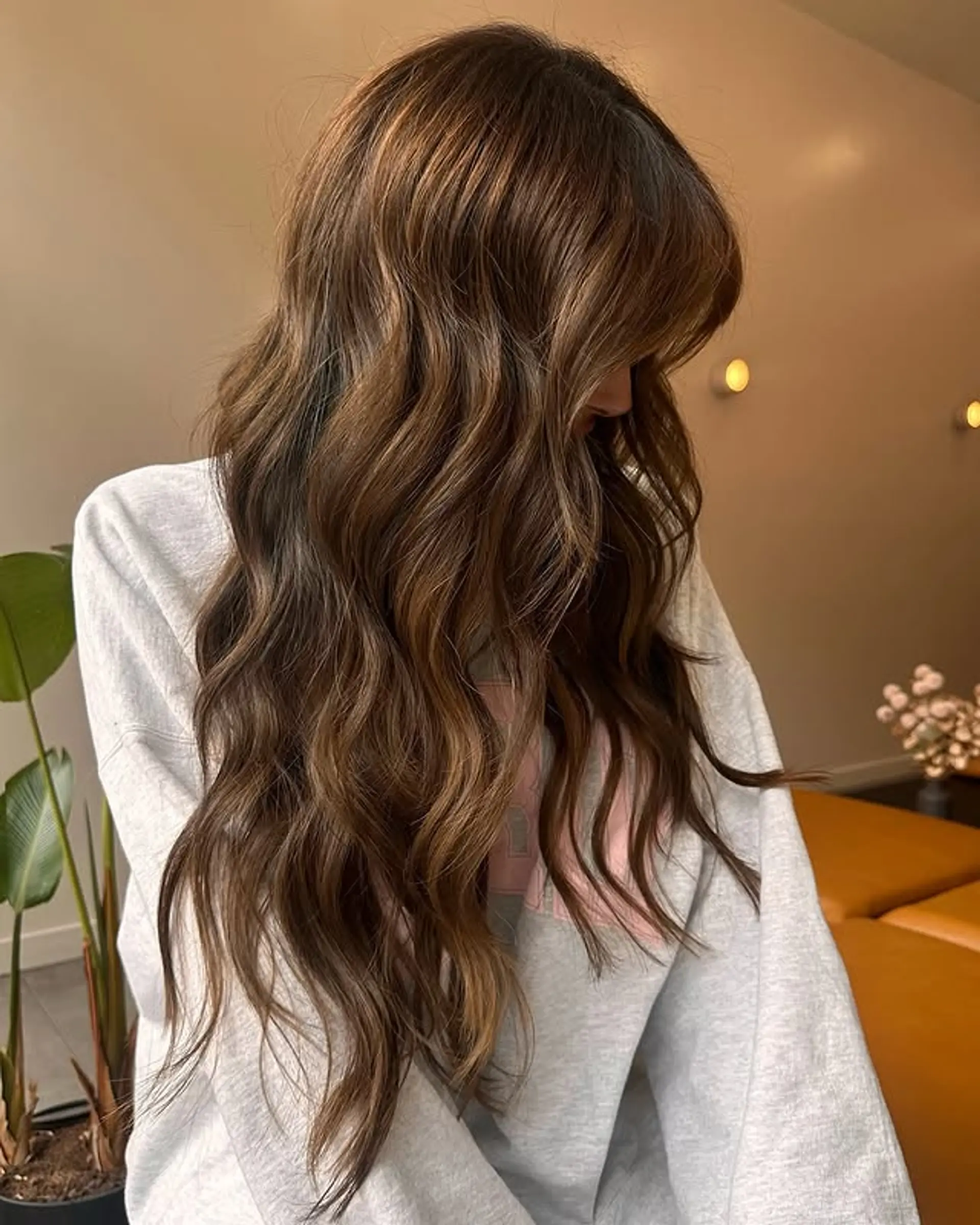 Long, wavy hair with layered cut and curtain bangs. Features a bronze balayage on a brunette base. 1