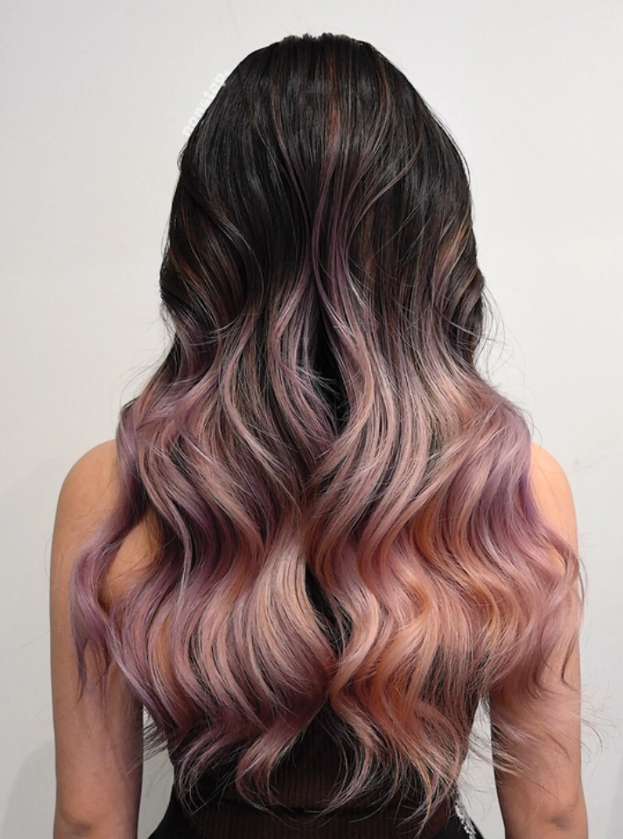 The hairstyle features long, wavy hair transitioning from natural black roots to a mix of rose gold and lavender towards the ends. The waves are loose and voluminous, with subtle balayage highlights adding depth and dimension.