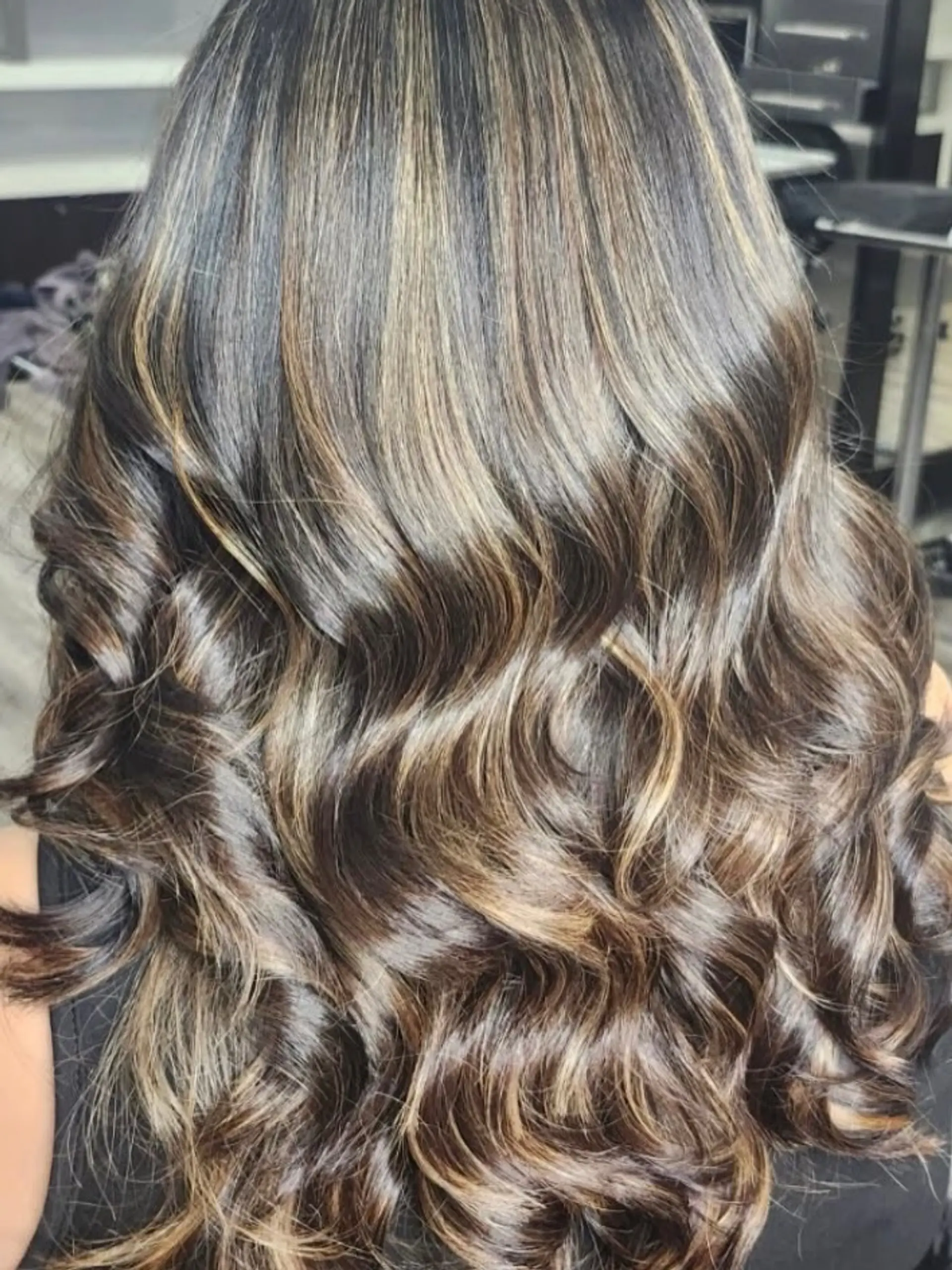 Long dark brown hair enhanced with dimensional caramel highlights and styled into shiny, cascading waves. 1