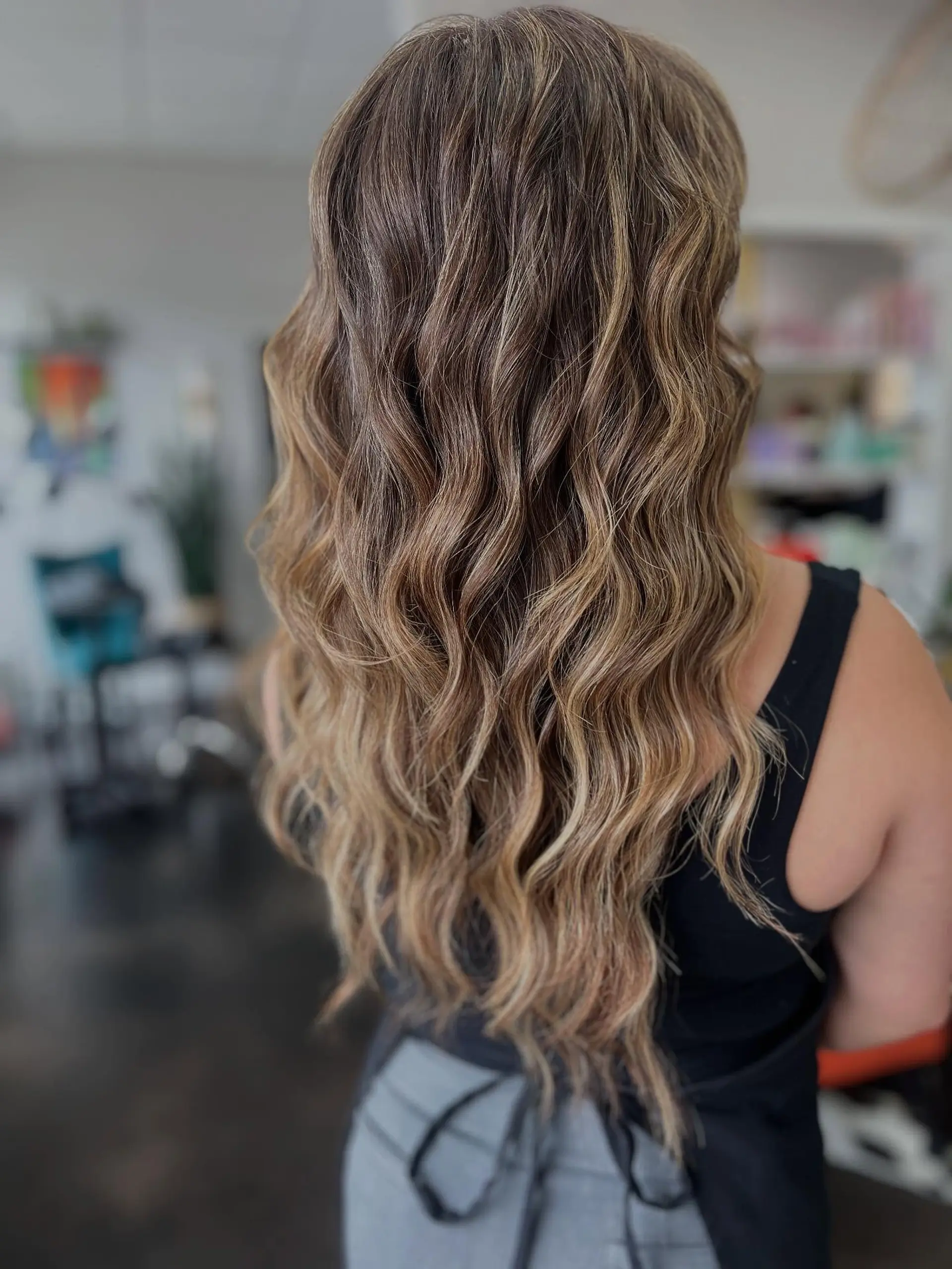 Long cascading beach waves featuring a seamless, blended balayage from warm brown roots to dimensional honey blonde tones. 1