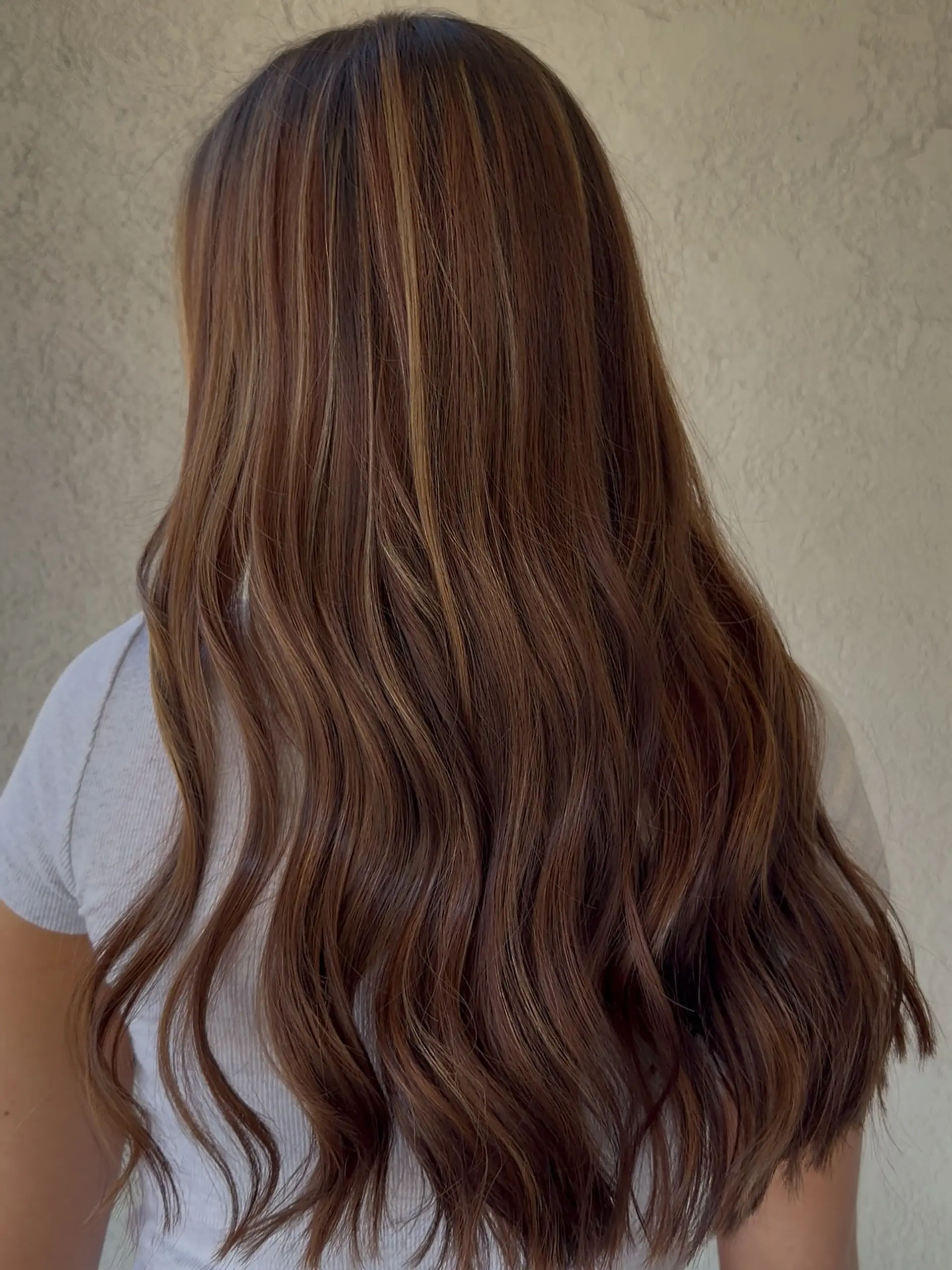 Long warm brunette hair styled in soft, loose waves featuring dimensional balayage highlights throughout. 1
