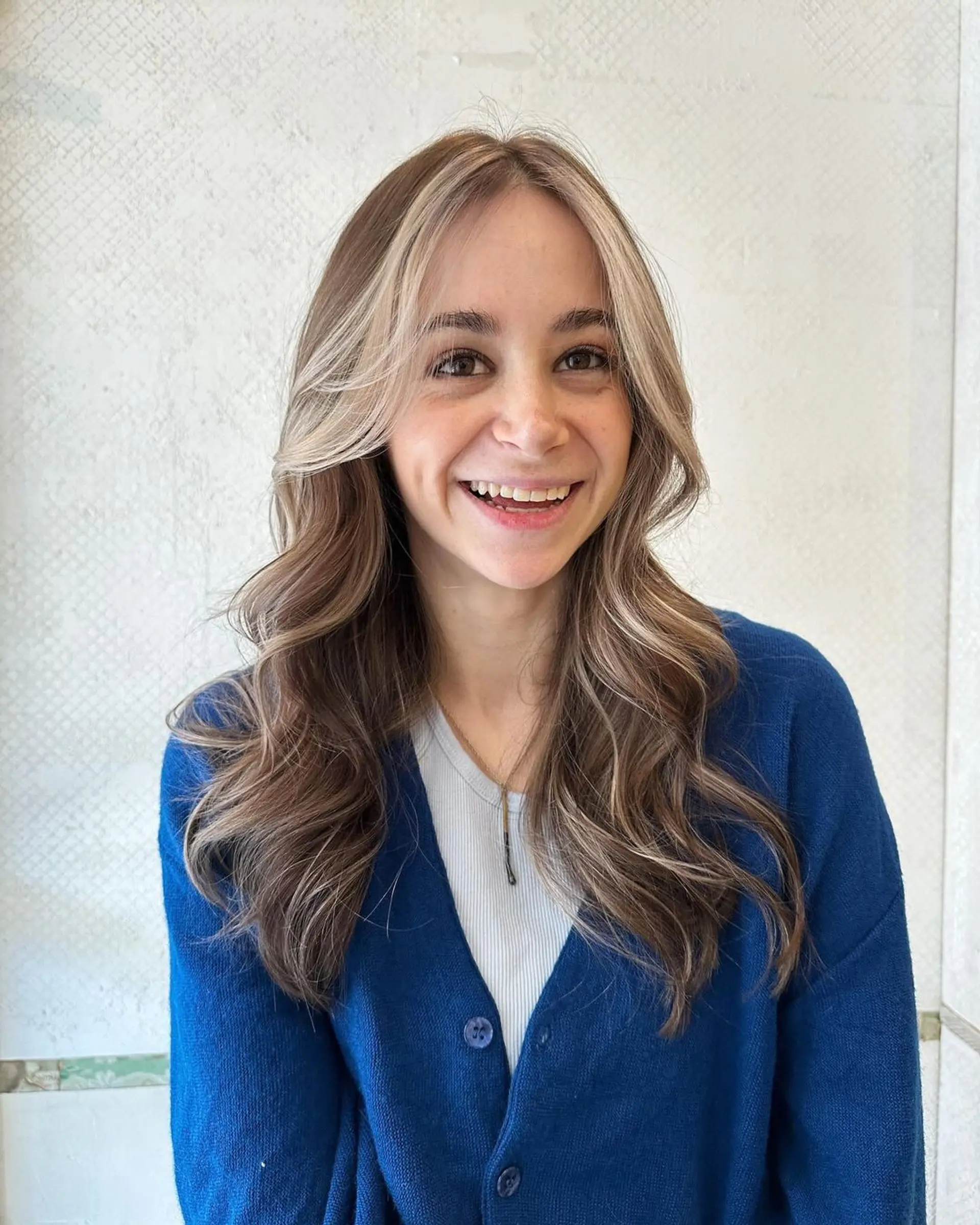 The hairstyle features long, wavy tresses with a blend of light brown and blonde colors, highlighted softly around the face. The layers add dimension and movement to the glossy waves. 1