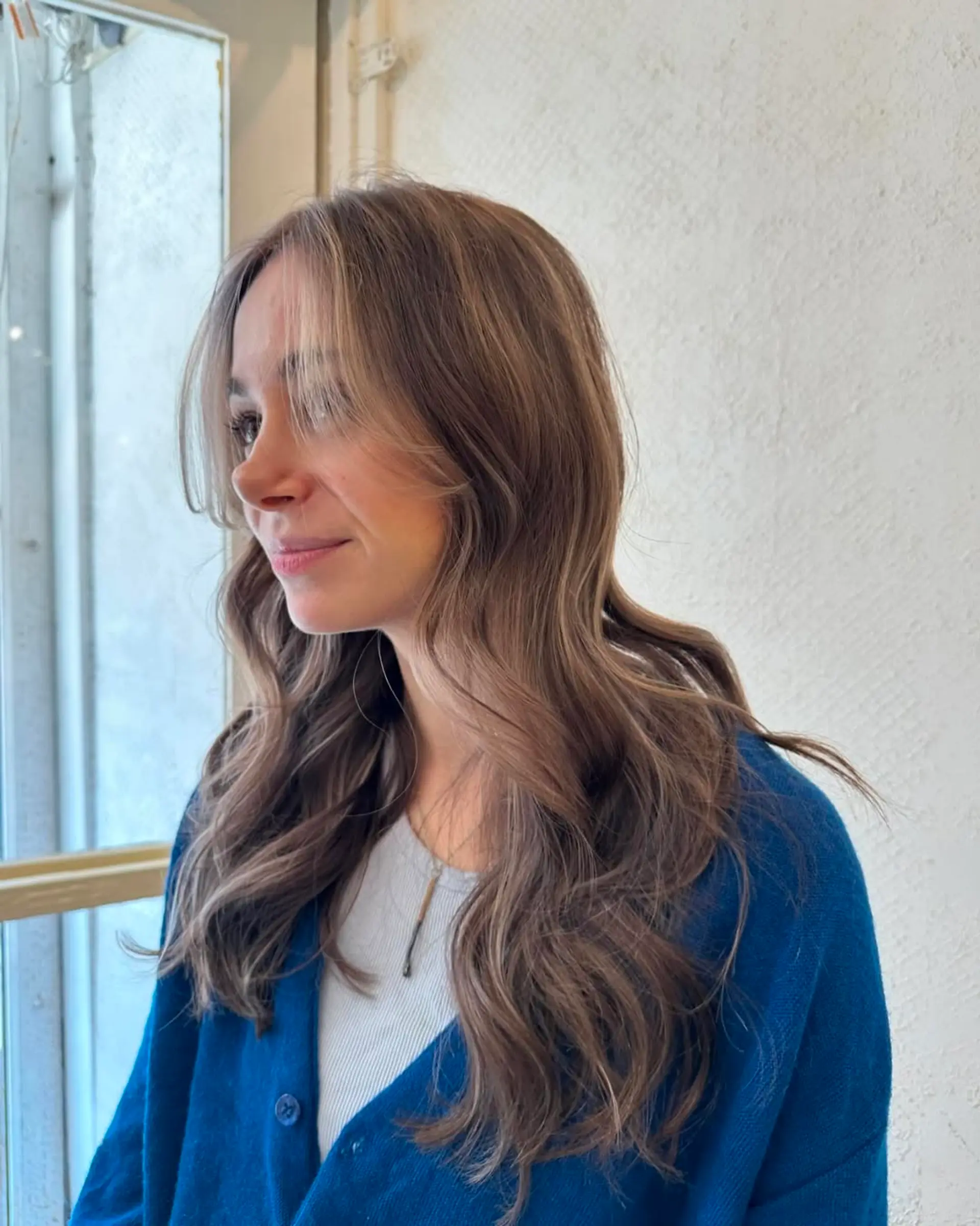 The hairstyle features long, wavy tresses with a blend of light brown and blonde colors, highlighted softly around the face. The layers add dimension and movement to the glossy waves. 2