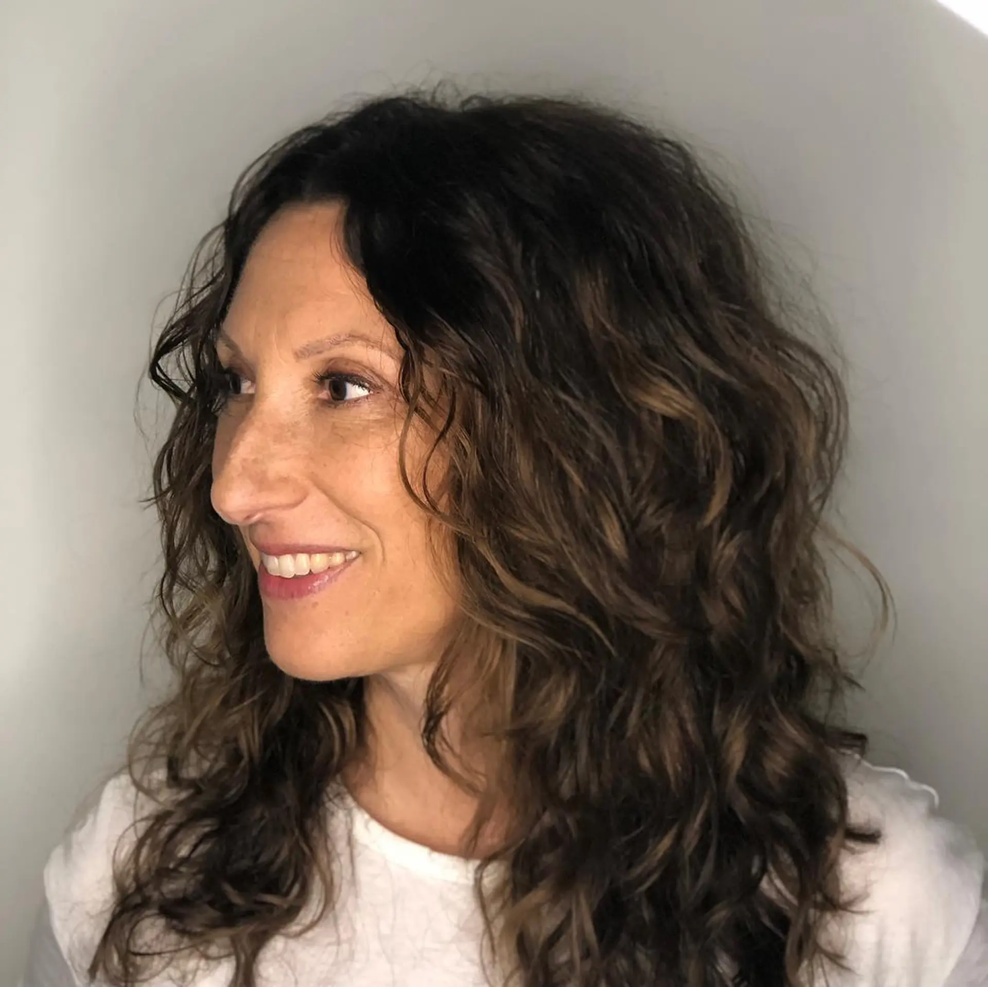 Medium-length layered haircut enhancing natural curls, featuring brunette tones with soft warm highlights. 1