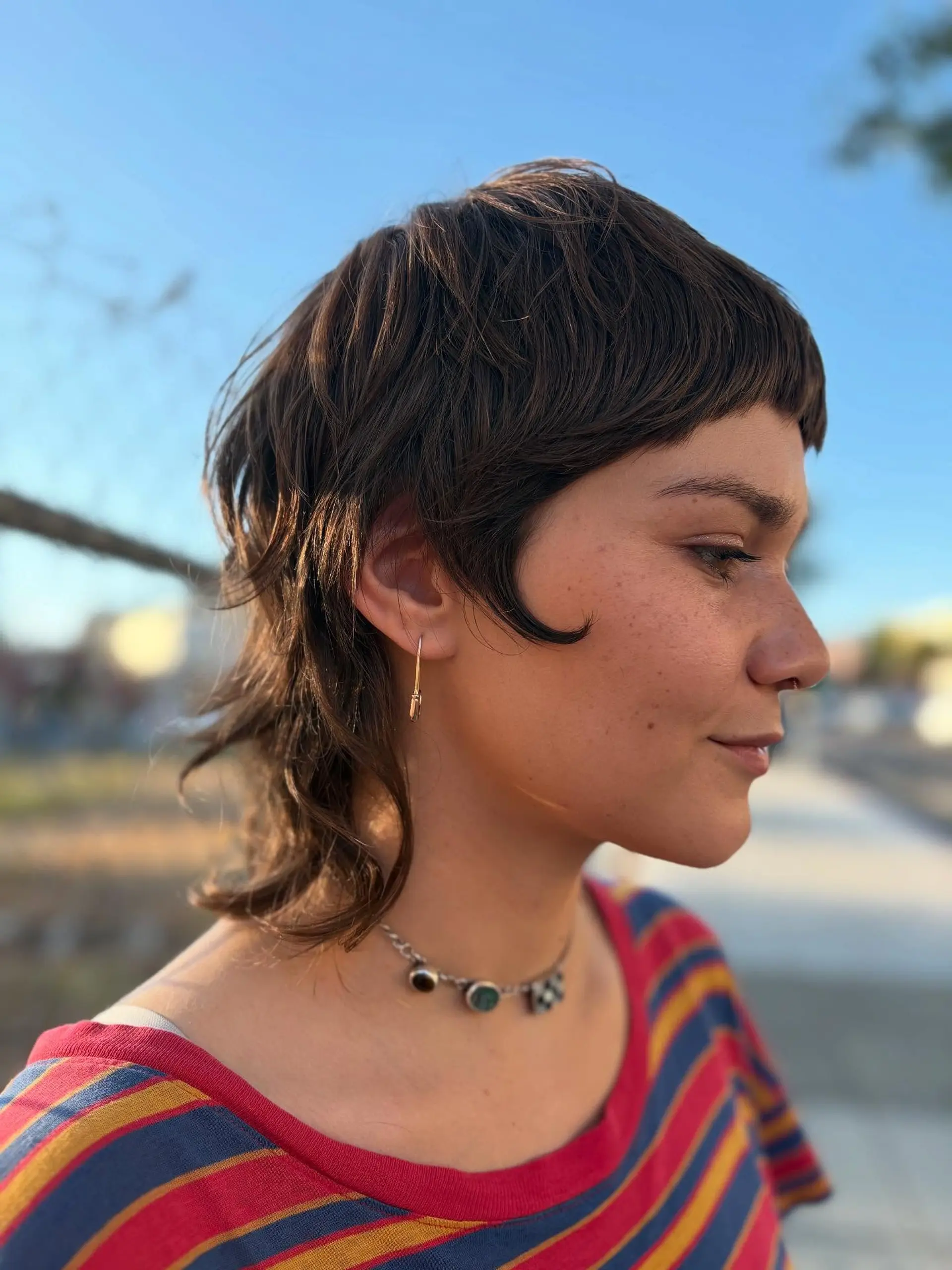 A modern razor-cut mullet featuring blunt micro bangs, forward-curving sideburn tendrils, and textured wavy length at the nape. 1