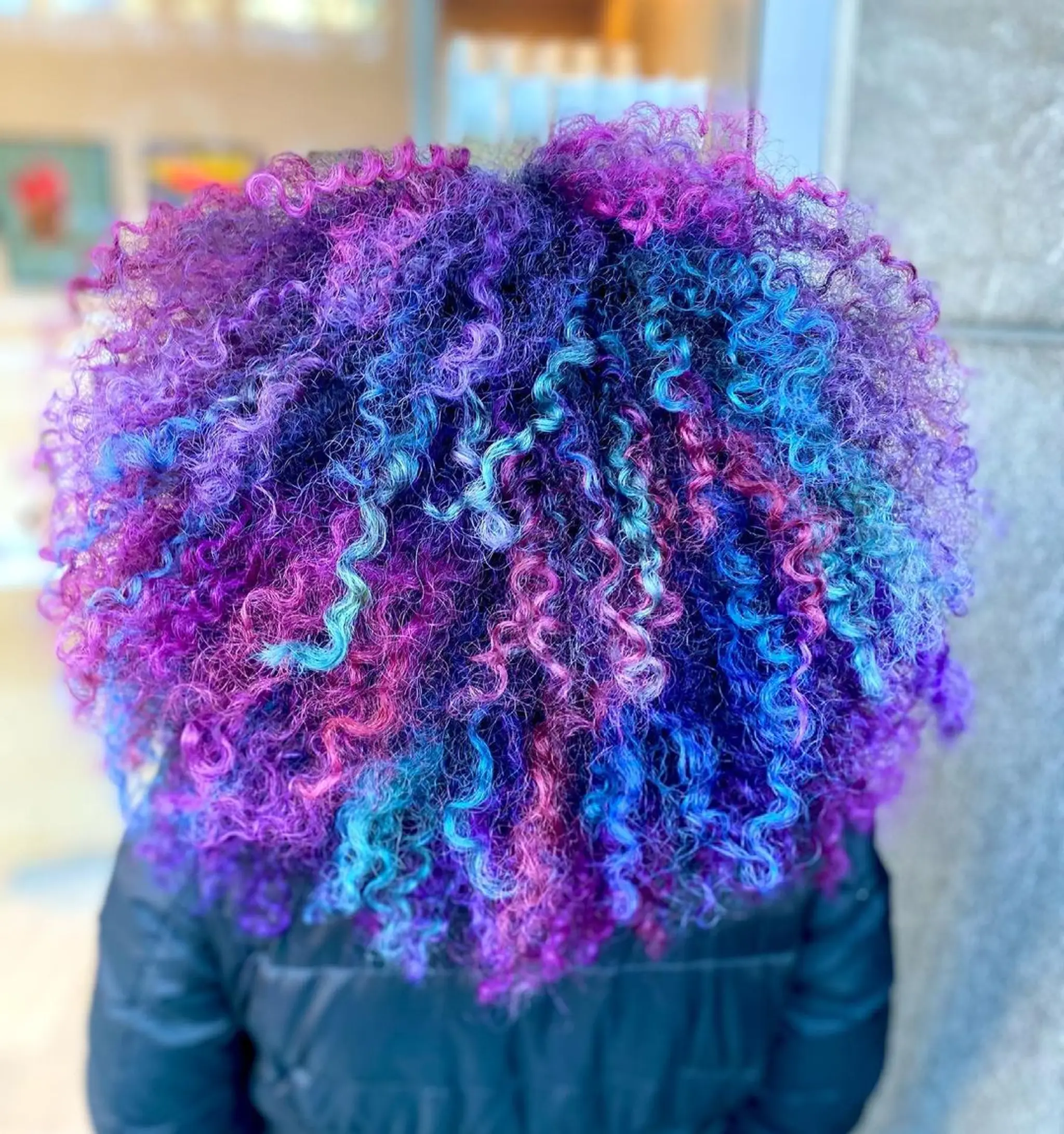Medium-length, curly hair in vibrant hues of purple, blue, pink, and teal, styled into a voluminous afro.