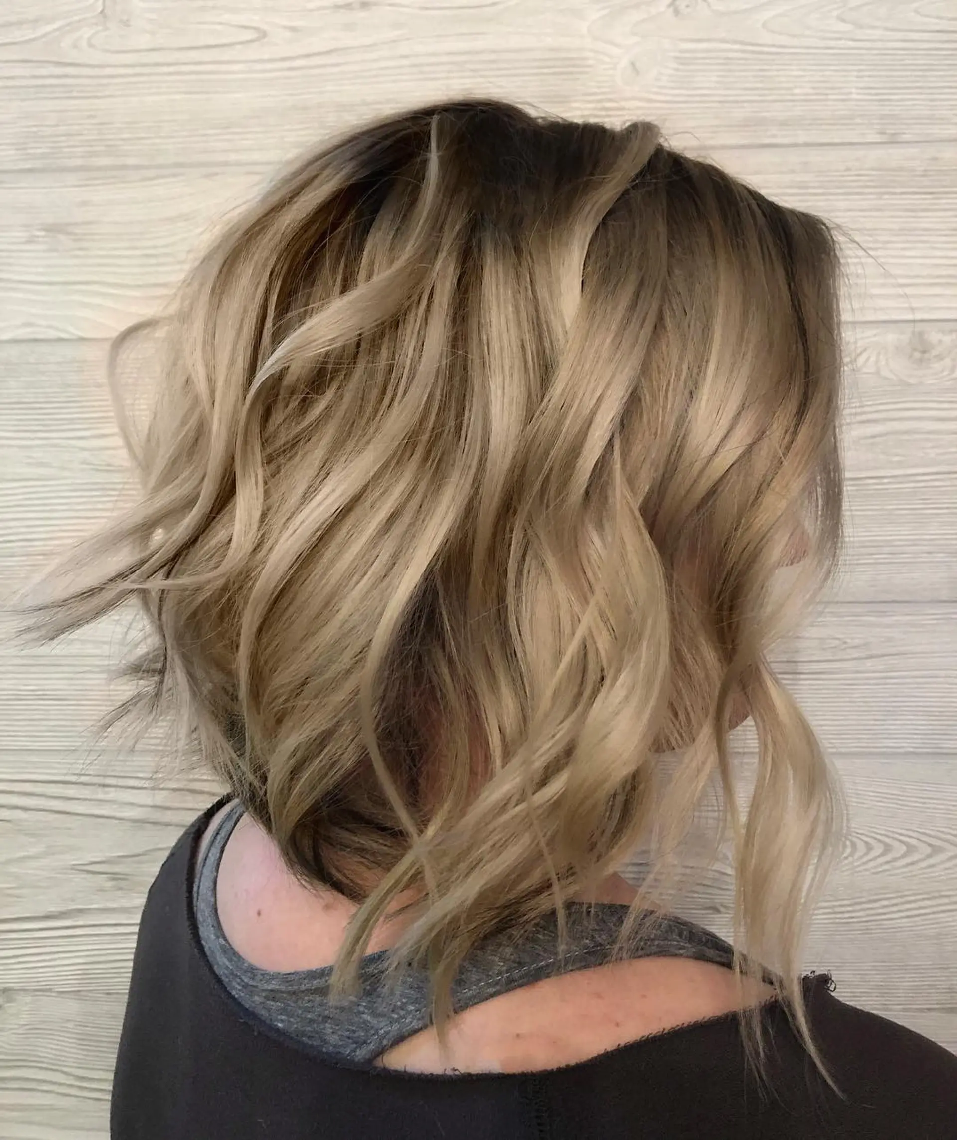 A textured, angled short bob styled with loose waves, featuring a dark shadowed root that smoothly melts into multi-dimensional warm and cool blonde tones. 1