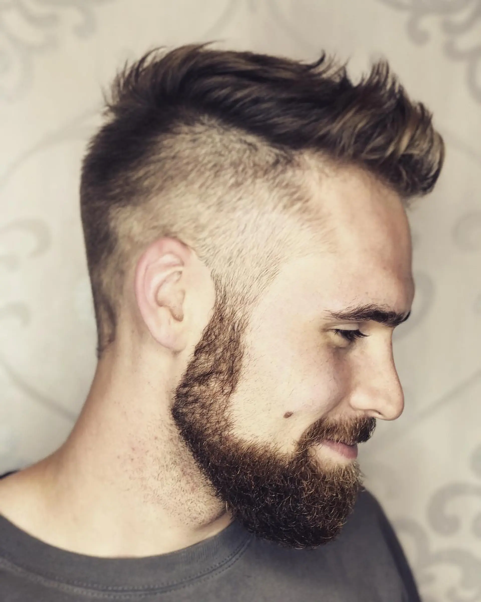 A modern men's undercut featuring high buzzed/faded sides and a textured, swept-up top, paired with a shaped, full beard. 1
