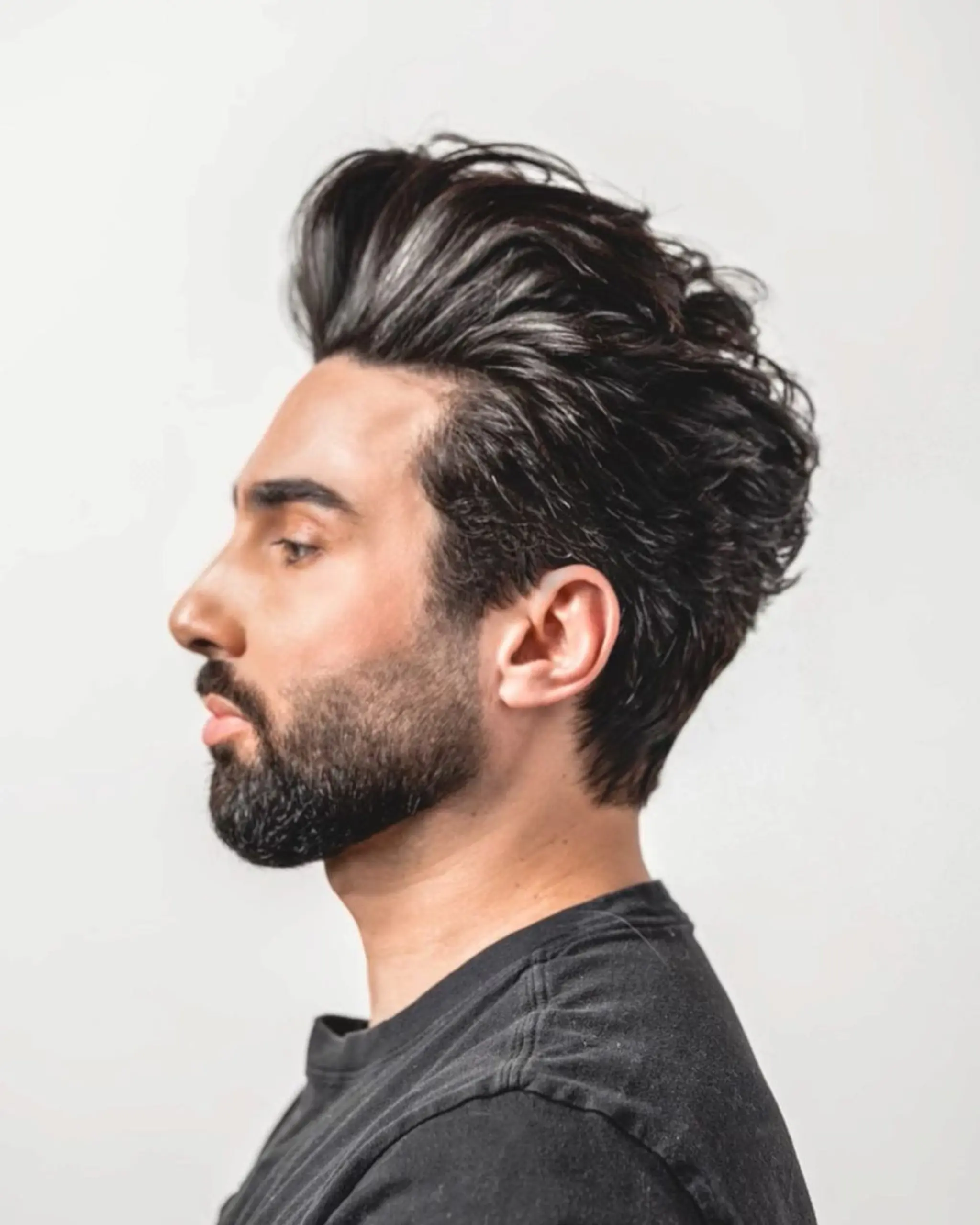 A voluminous pompadour with high waves at the front, sleek sides, and a textured finish. Medium-length black wavy hair adds height and sophistication to the style. Cut by @robincapili