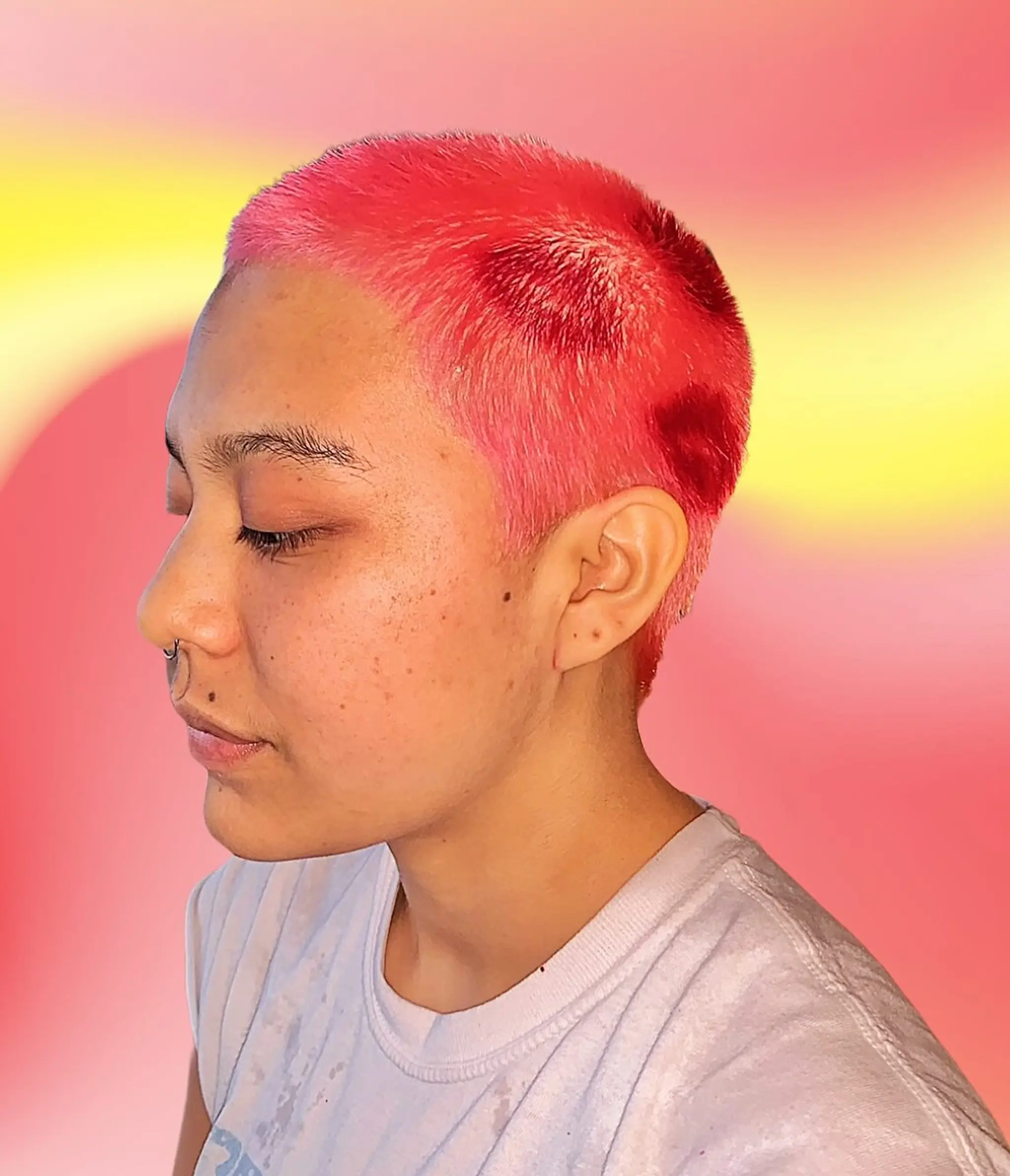 A very short, vibrant buzz cut featuring neon pink and red shades with a gradient effect.