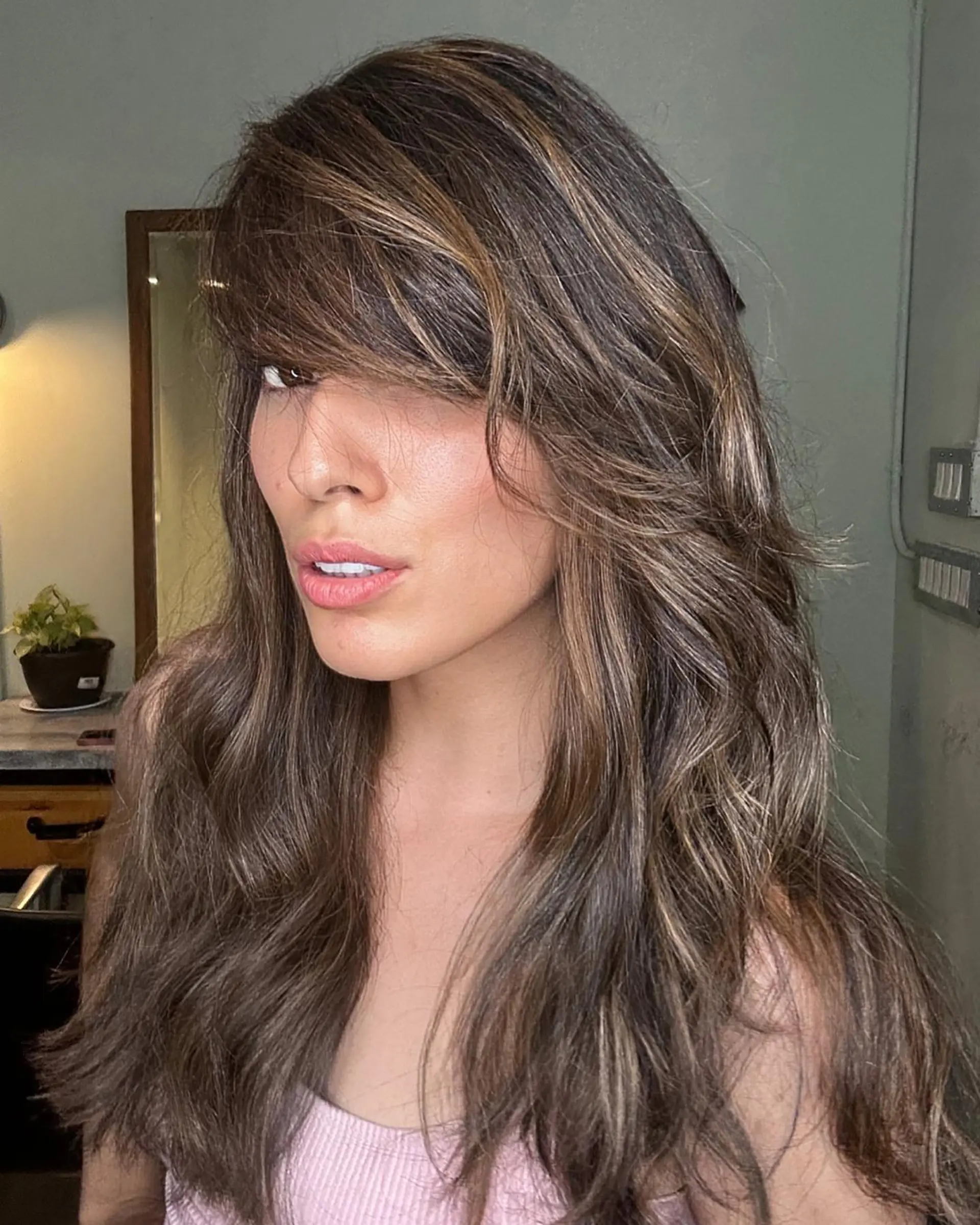 Long, layered brunette hair featuring warm caramel highlights and styled with voluminous side-swept bangs. 1
