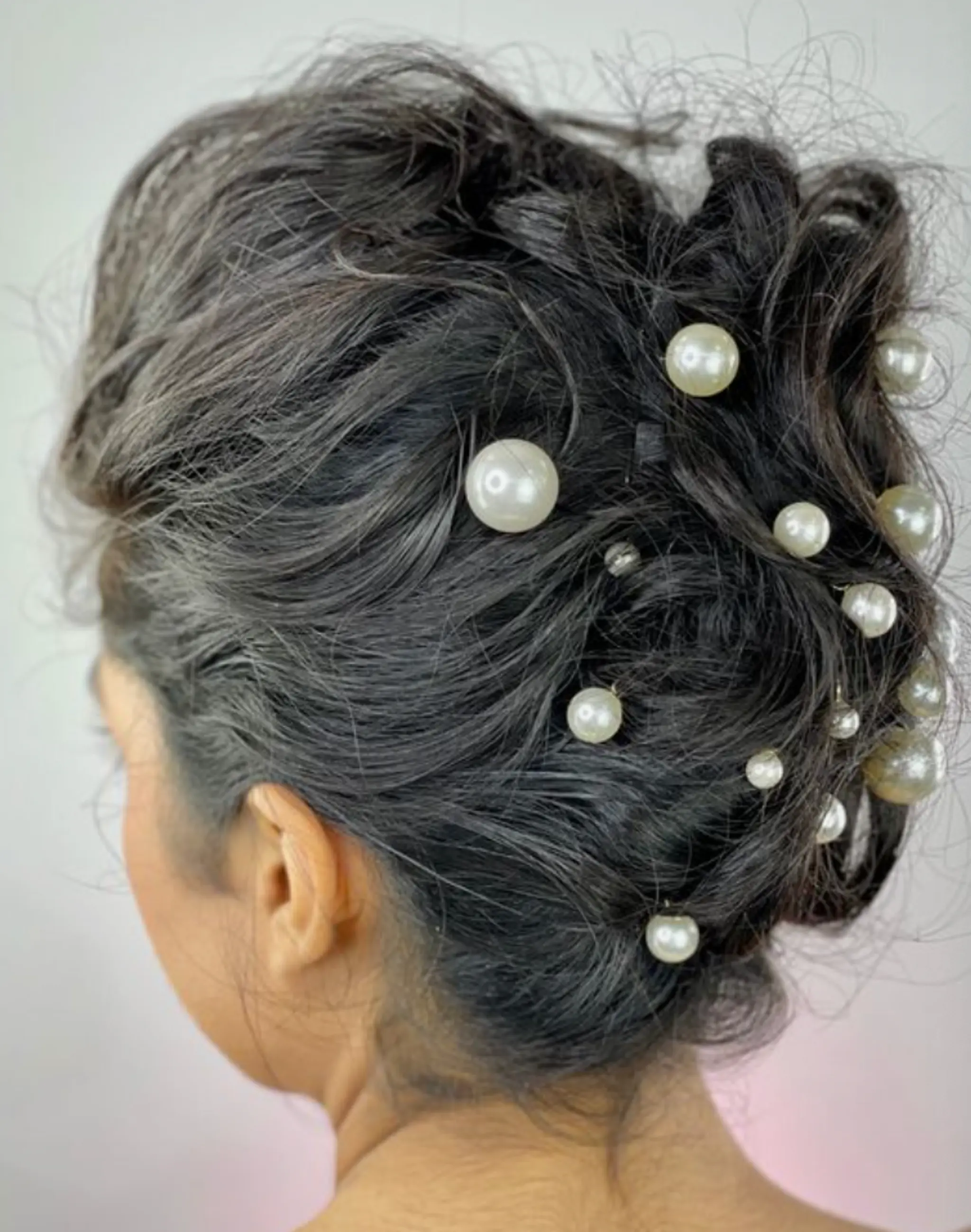 An elaborate updo with medium-length wavy black hair intricately twisted into an elegant bun, adorned with scattered pearlescent beads.