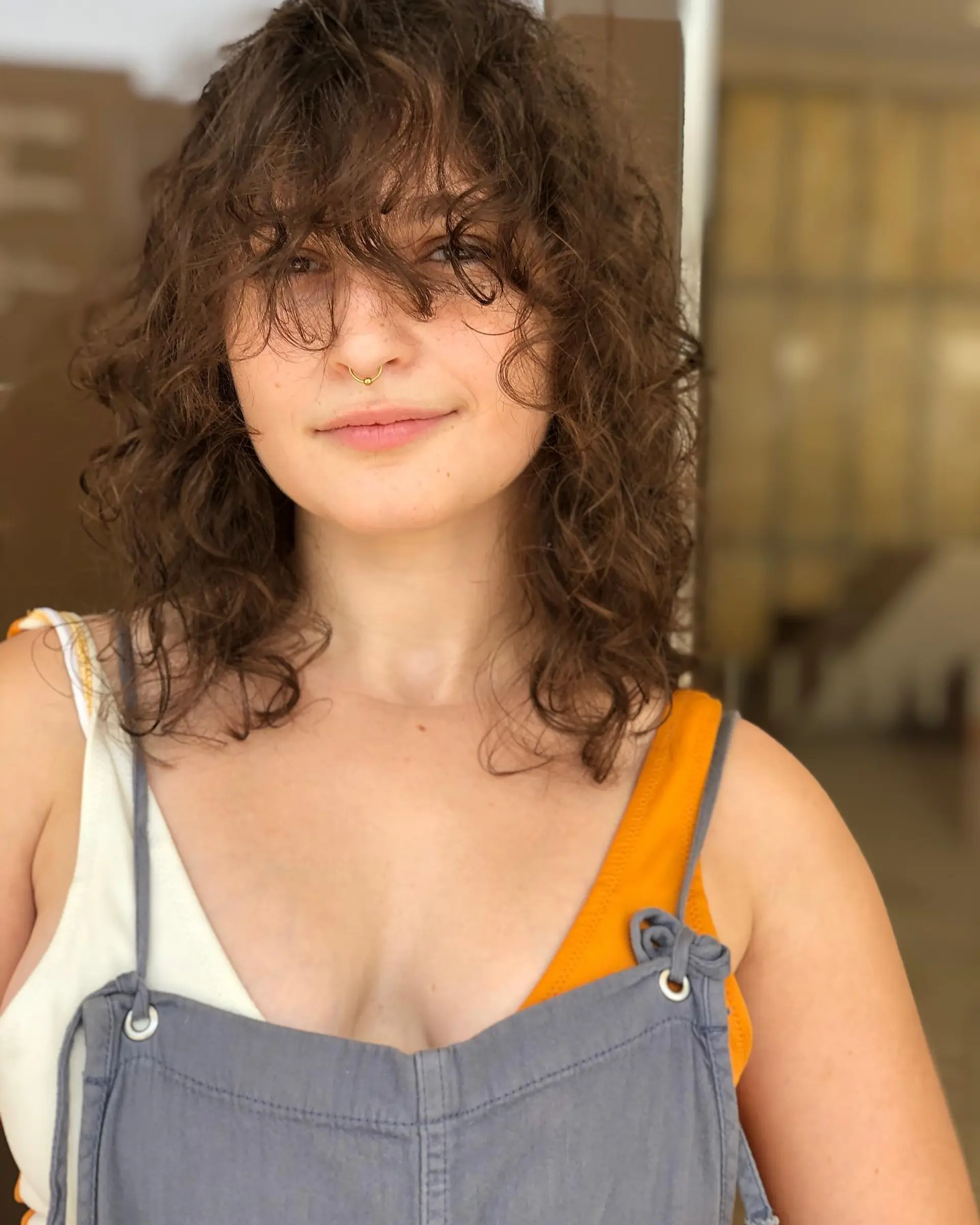 A medium-length curly shag cut featuring full, wispy bangs falling over the eyes and textured curly layers. 1